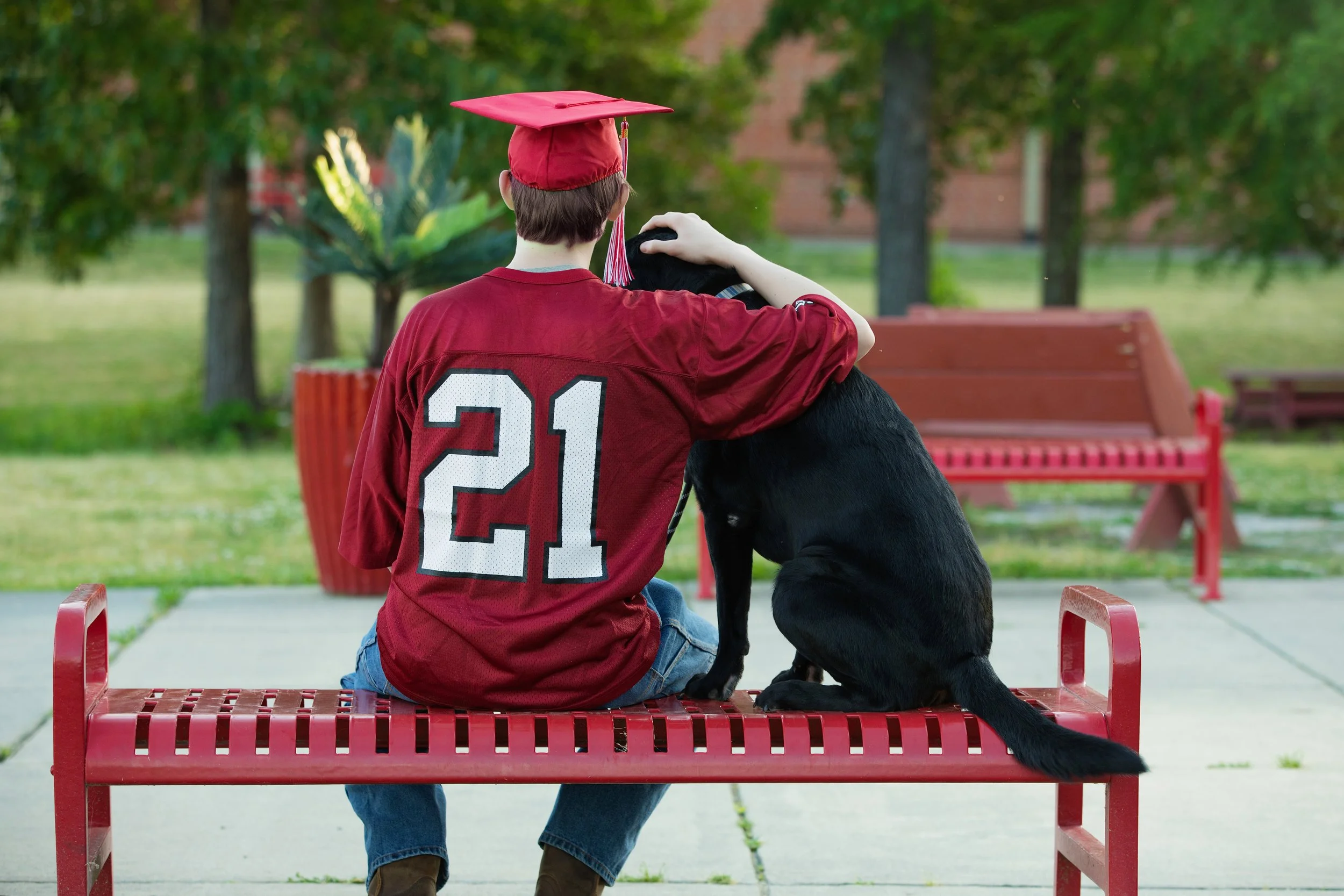 Highschool senior boy wearing a football jersey and his graduation cap sitting on a bench with his dog.