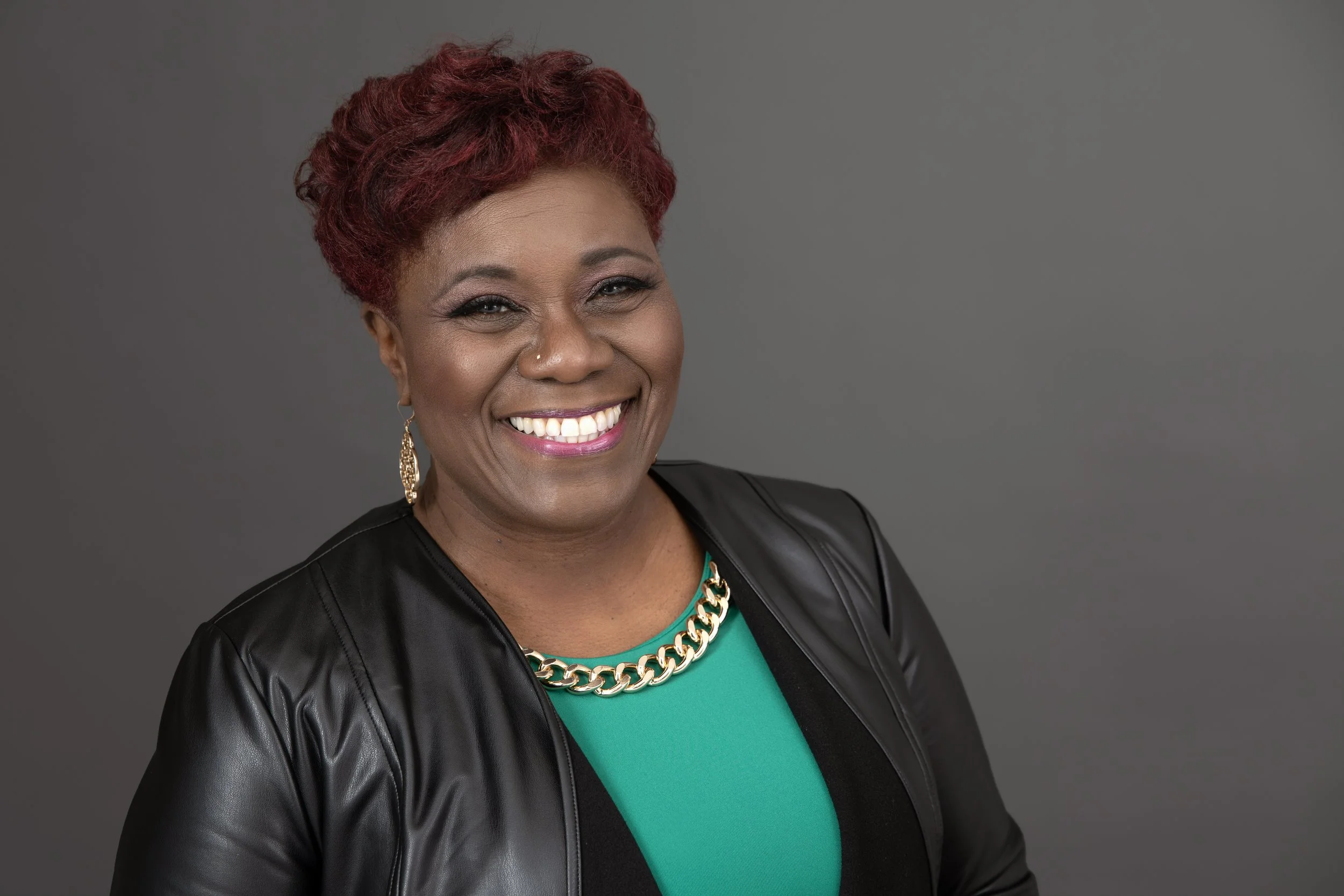 A smiling woman with short, curly red hair, wearing a black leather jacket, a green top, a gold chain necklace, and gold earrings, against a solid gray background.
