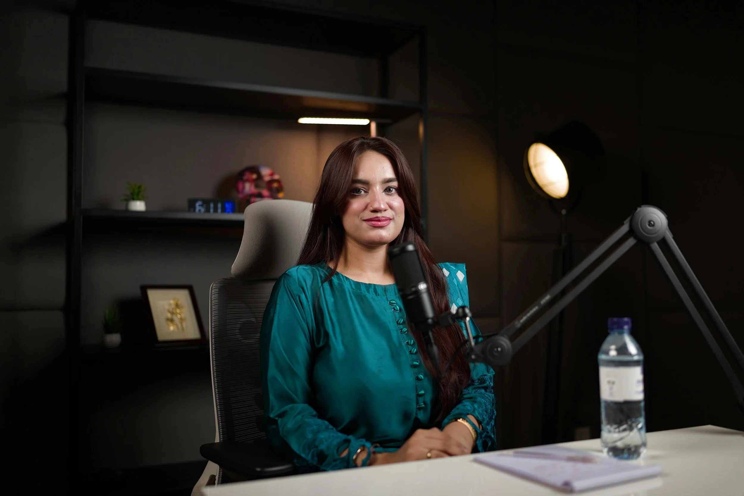A woman with long brown hair, wearing a teal blouse, sitting at a desk with a microphone, water bottle, and notebooks in front of her, in a dark office studio setting.