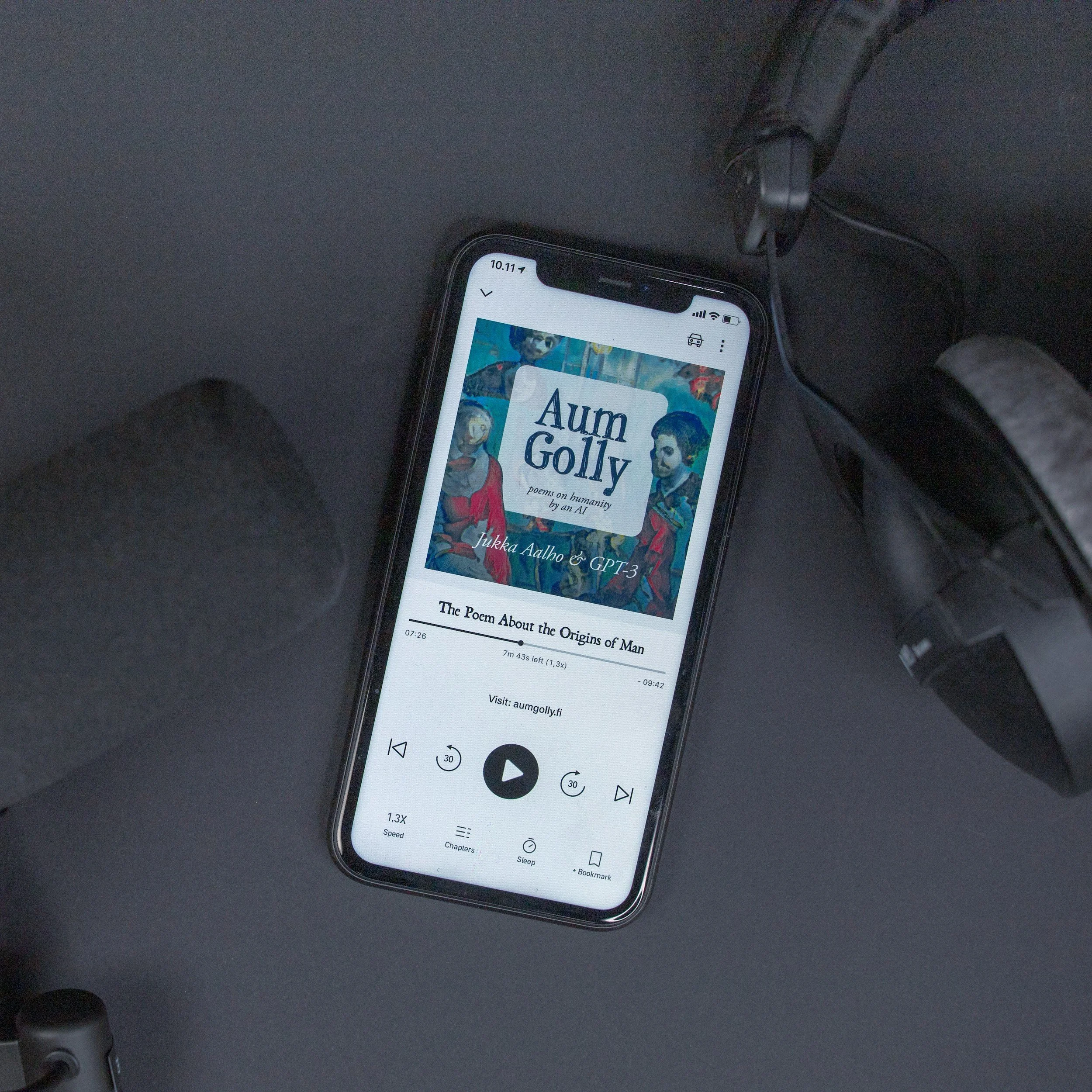 A smartphone resting on a dark surface playing an audiobook www studios com titled 'Aum Golly' with a colorful cover art featuring artistic illustrations of people, connected to a pair of black headphones.
