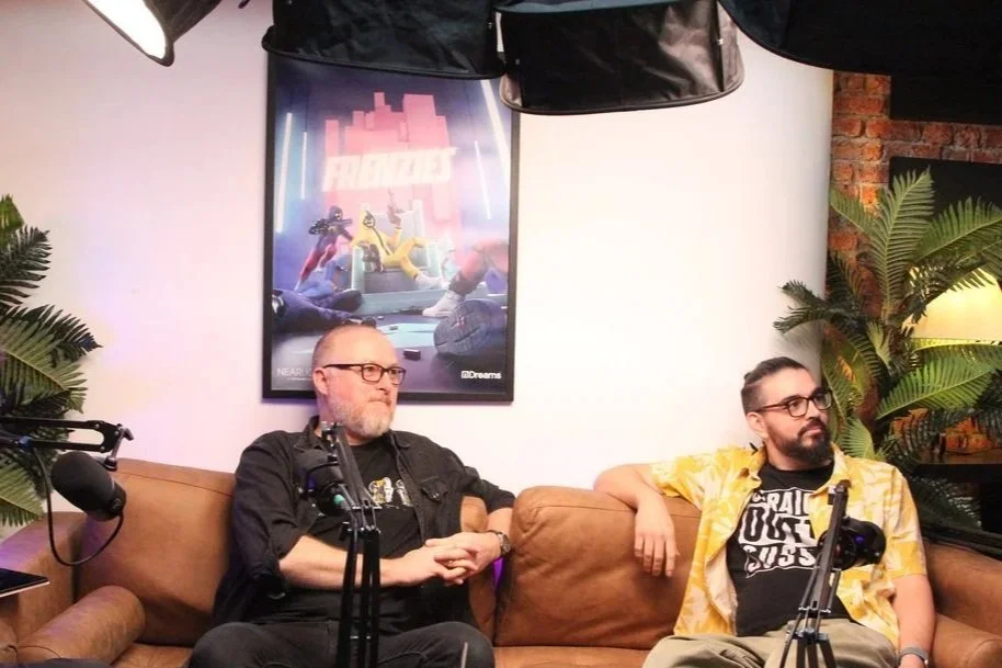 Podcast recording setup with three men seated on a couch, microphones, a poster on the wall, and plants in the background.