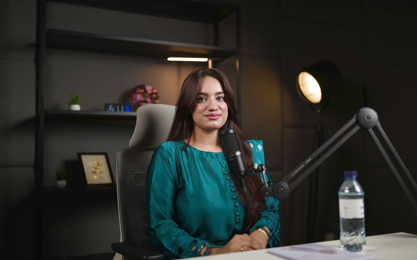 A woman with long brown hair sitting at a desk in a podcast studio London creators trust, with a microphone in front of her, a water bottle on the desk, and a black bookshelf behind her.