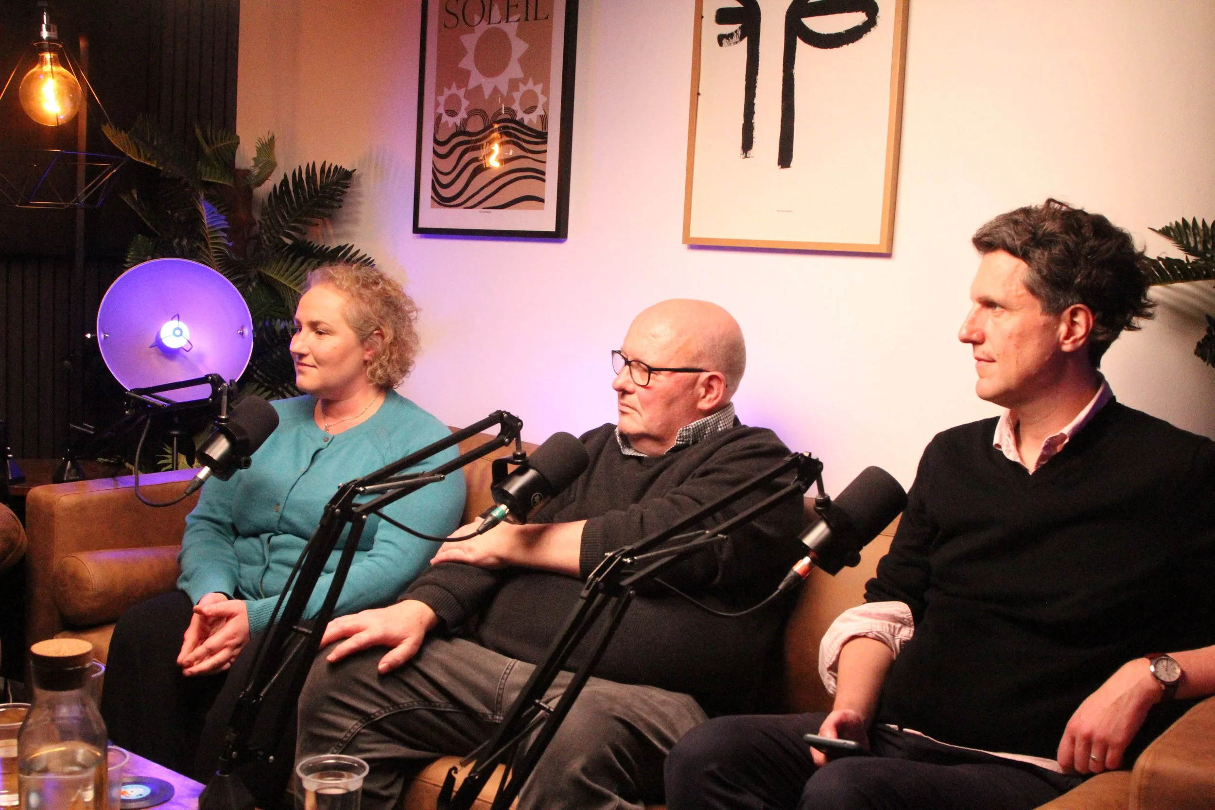Three people sitting on a sofa during a podcast recording, with microphones in front of them. There is a woman on the left, a man with glasses in the middle, and another man on the right. The background features framed artwork and plants.