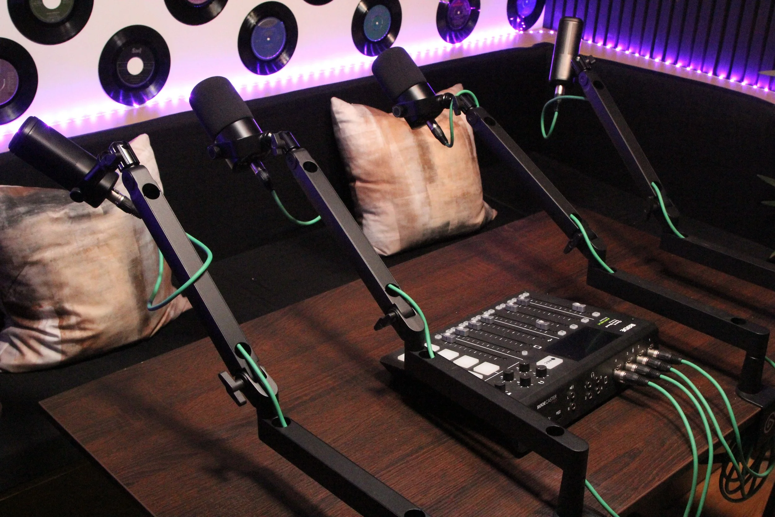 Podcast recording studio with four microphones on adjustable arms hanging over a wooden table, a mixing console, and pillows on a black couch, decorated with vinyl records and purple LED lighting.