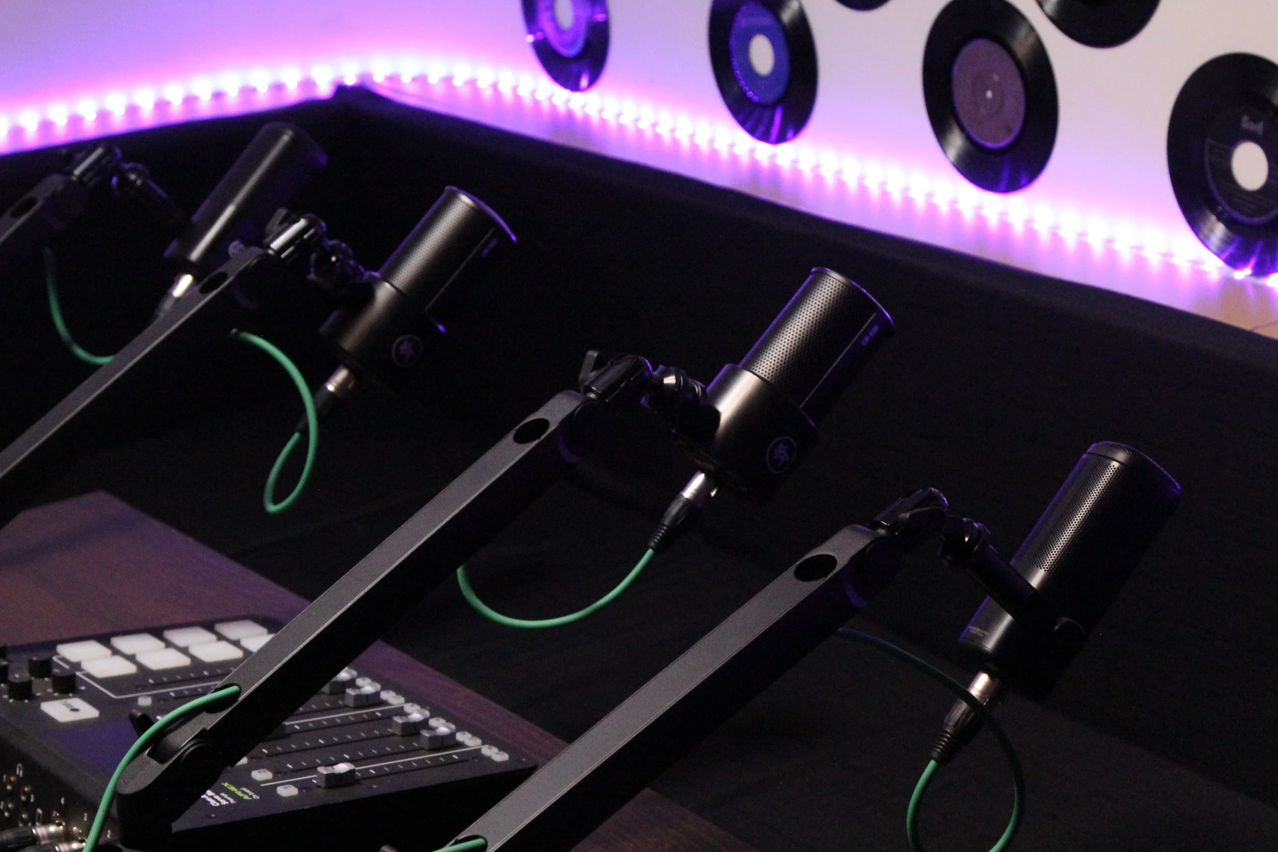 Podcast recording studio setup with three microphones mounted on adjustable booms, a sound mixer, and a wall decorated with vinyl records illuminated by purple LED lights.