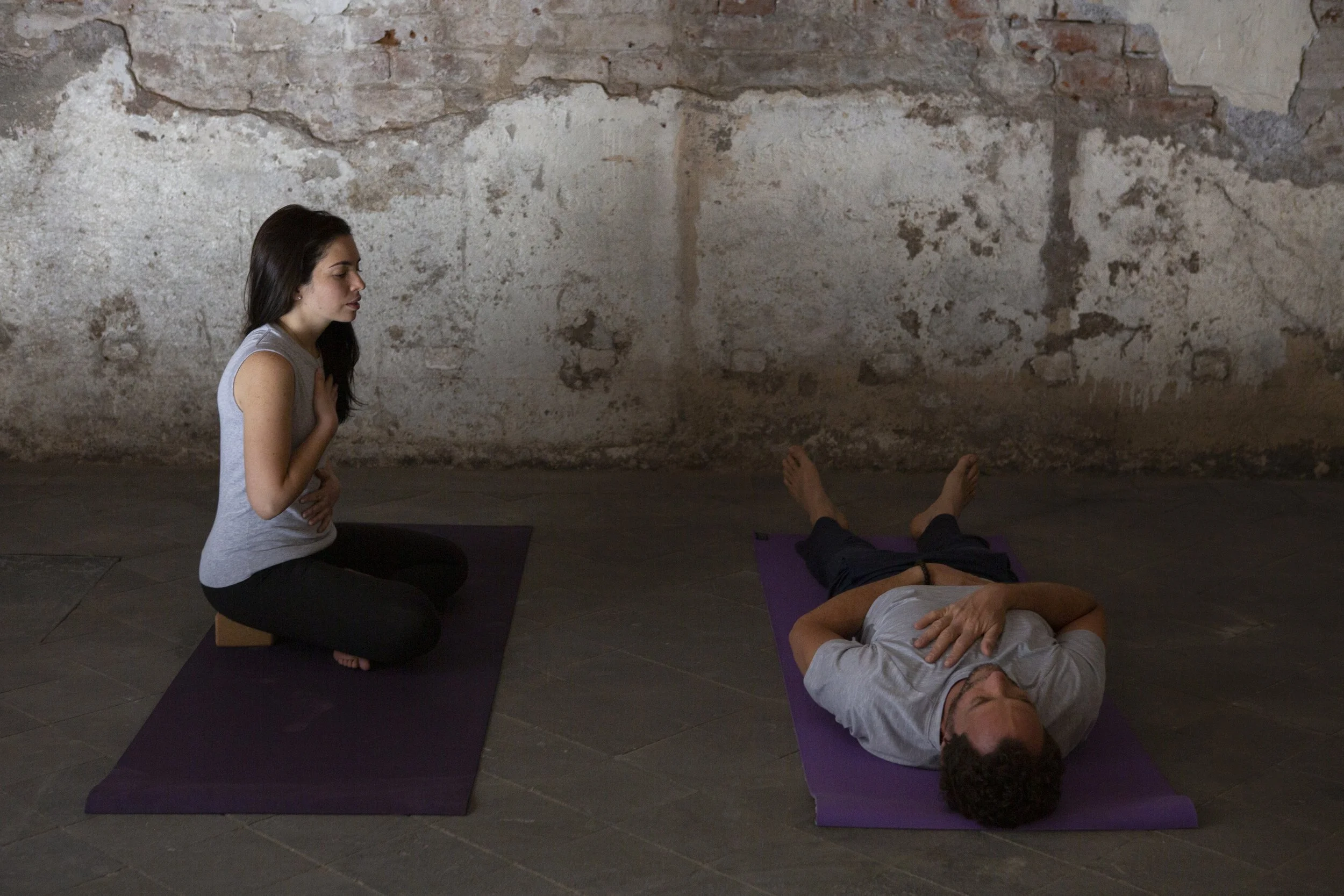 Una mujer haciendo yoga en una colchoneta morada frente a un hombre acostado sobre una colchoneta morada en un suelo de piedra y un fondo de muro de ladrillo y yeso