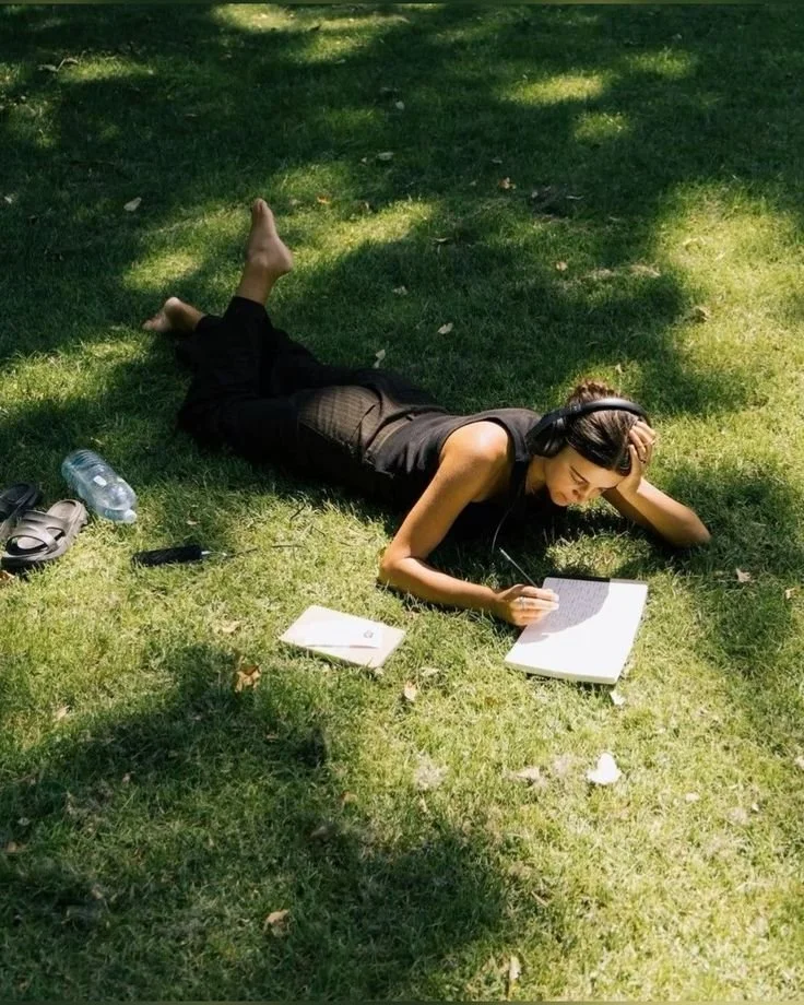 Mujer recostada en el pasto leyendo y escuchando música con audífonos, rodeada de libros, botella de agua y objetos personales en un parque