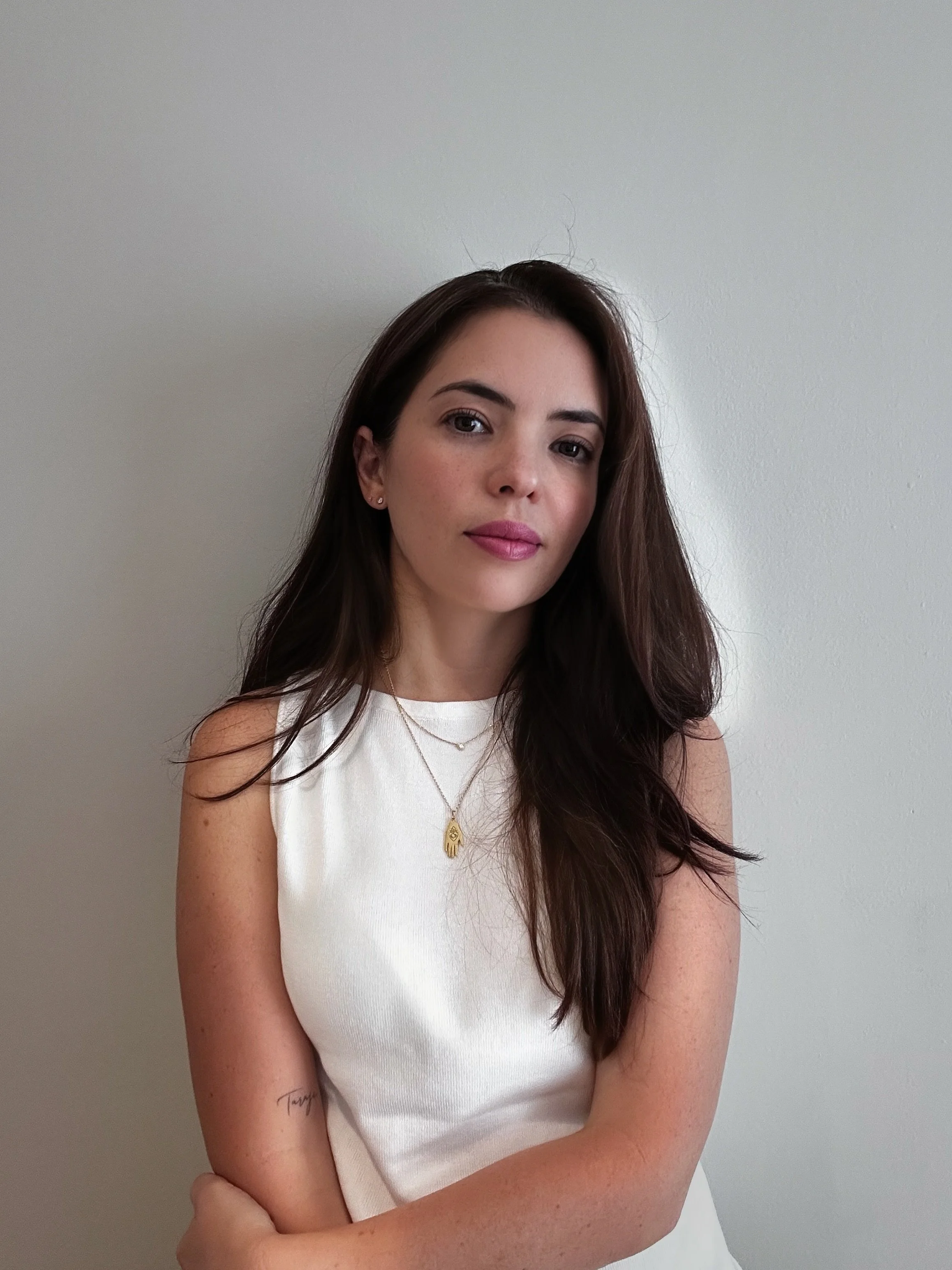 Retrato de una mujer con cabello largo y oscuro, usando blusa blanca y collar dorado, posando frente a una pared blanca.