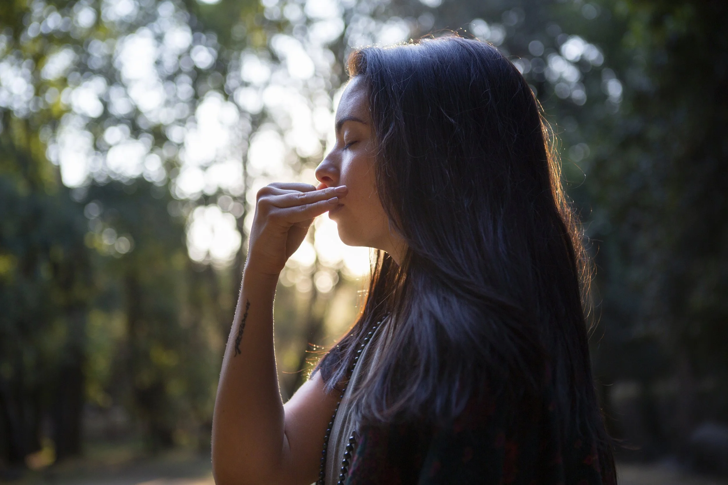 Mujer con cabello largo y oscuro, de perfil, con ojos cerrados, tocándose la nariz con la mano, en un entorno natural con árboles y luz solar en el fondo.