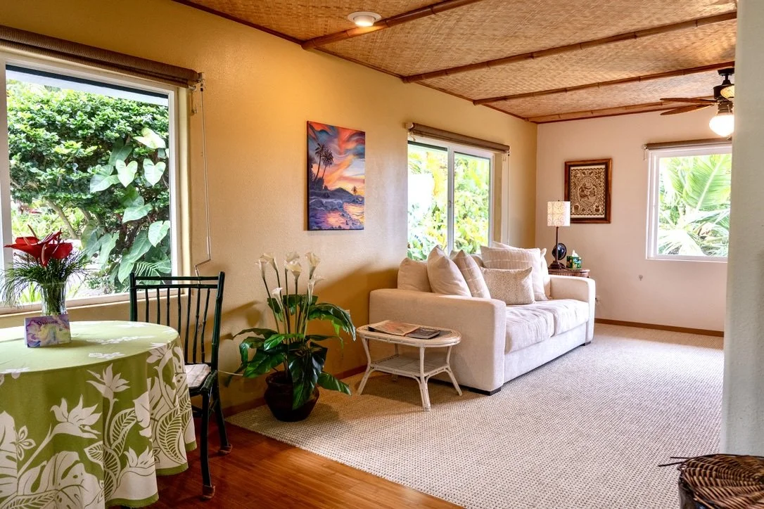 Living room with beige sofa, large windows with greenery outside, potted plant, round dining table with floral tablecloth and vase of flowers, artwork on walls, ceiling fan, and table lamp.