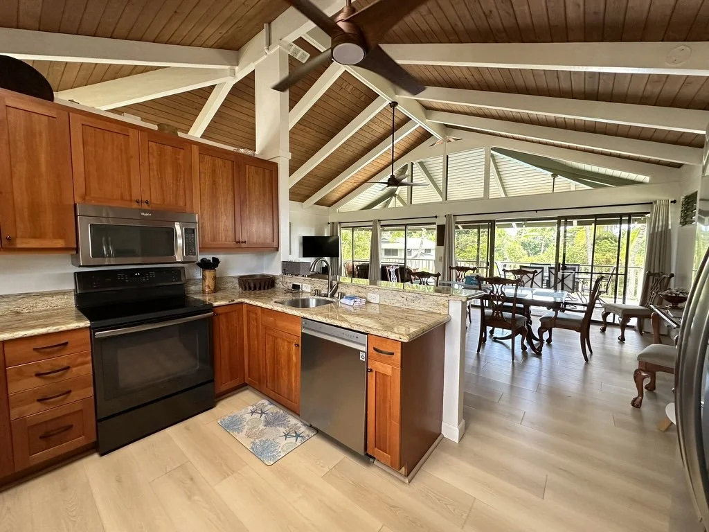 Open-concept kitchen and dining area with wooden cabinets, granite countertops, black appliances, and a vaulted ceiling with exposed beams and ceiling fans, leading to a sunlit dining space with large windows and outdoor views.