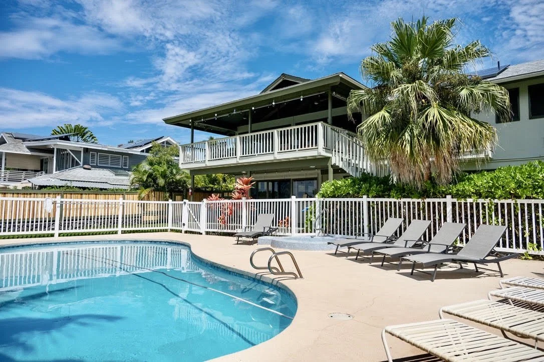 A backyard swimming pool area with lounge chairs, surrounded by a white fence, houses, a large palm tree, and a blue sky with some clouds.