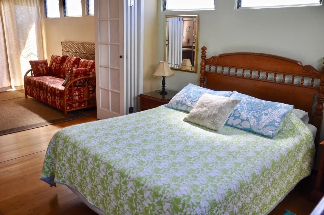 A bedroom with a wooden bed, pillows, a mirror, a nightstand with a lamp, and a sitting area with a floral-patterned sofa near a sliding door with curtains.