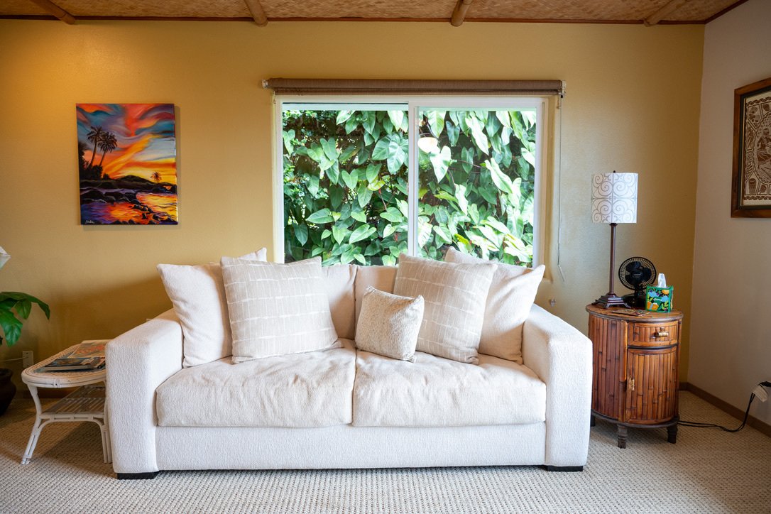 A living room with a white sofa, beige pillows, and a window showing green foliage outside.