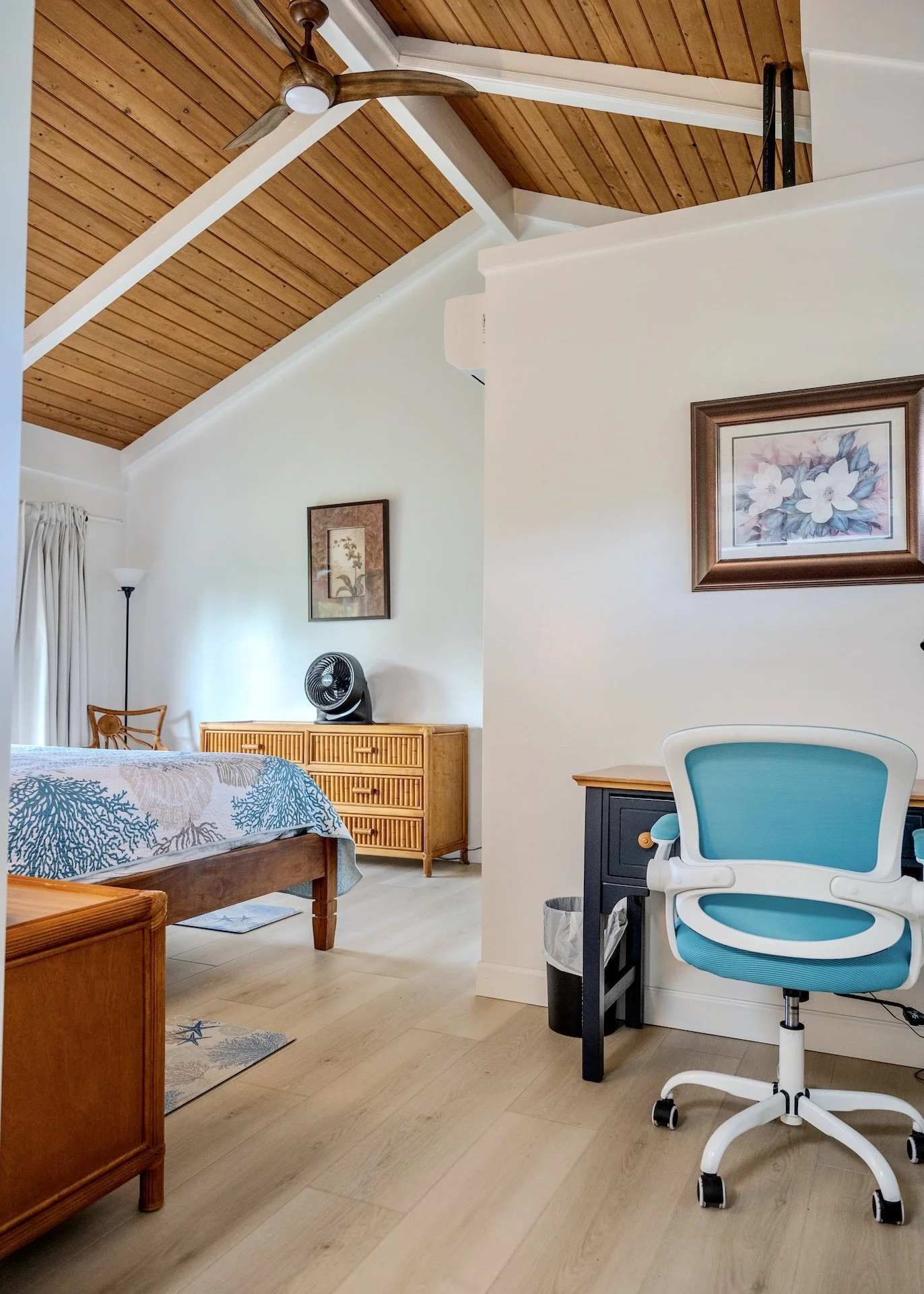 A room with a wooden ceiling, a bed with a coral-patterned bedspread, a wooden dresser with a small black fan on top, framed floral artwork on the walls, a white office chair with blue cushion, a small black desk, and a black trash can.