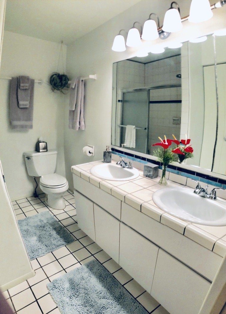 Bathroom with double sinks, mirror, toilet, towel racks, and shower area. Decor includes a vase with red flowers on the sink countertop and blue bath mats on the tiled floor.