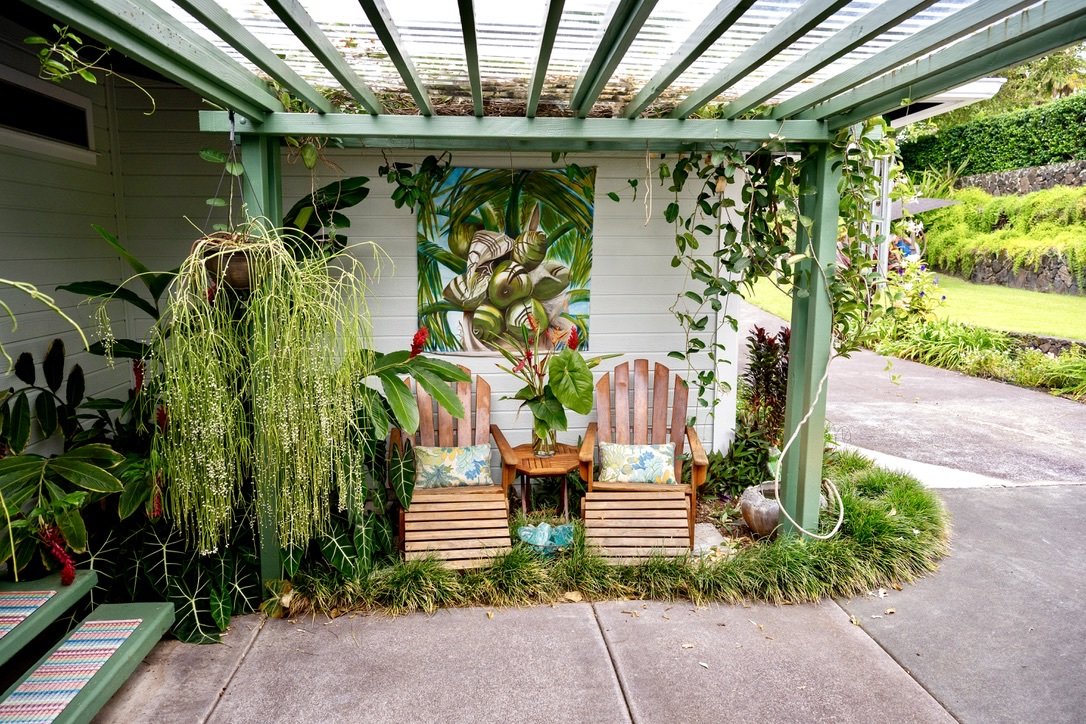 Outdoor patio with two wooden chairs, a small table with a potted plant, colorful garden painting, and lush green plants on a porch with a green pergola, paved walkway, and landscaped yard.
