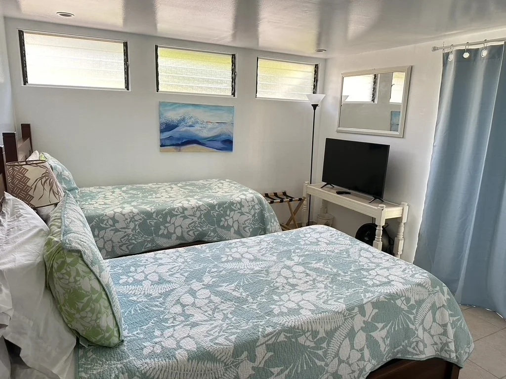 Twin beds with patterned quilts and pillows in a bright hotel room with a TV, lamp, and window blinds.
