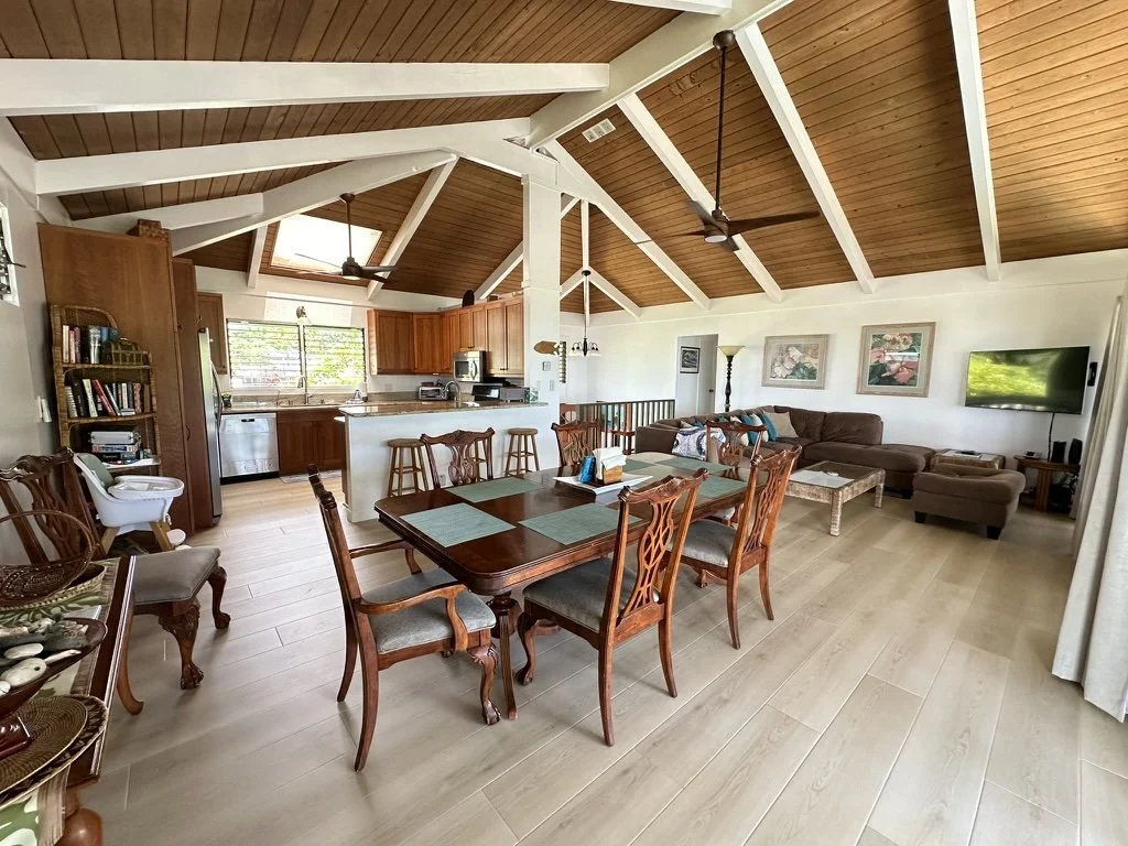Open-concept living room and kitchen with wooden vaulted ceiling, dining table with six chairs, brown sectional sofa, flat-screen TV, and large windows