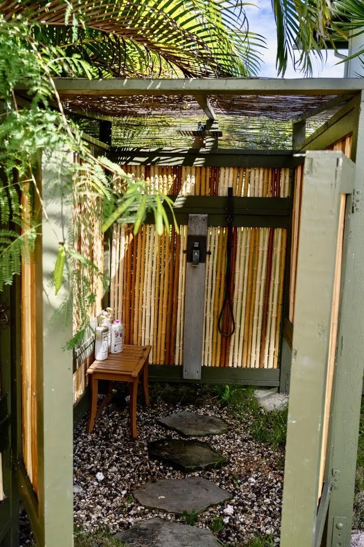 An outdoor shower with bamboo fencing, a small wooden bench with bottles of shower products, stepping stones on gravel, surrounded by lush green tropical plants and palm leaves overhead.