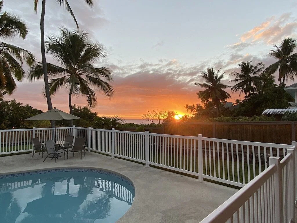View of a backyard swimming pool at sunset with palm trees, outdoor table with chairs and an umbrella, wooden fence, and a colorful sky with clouds.