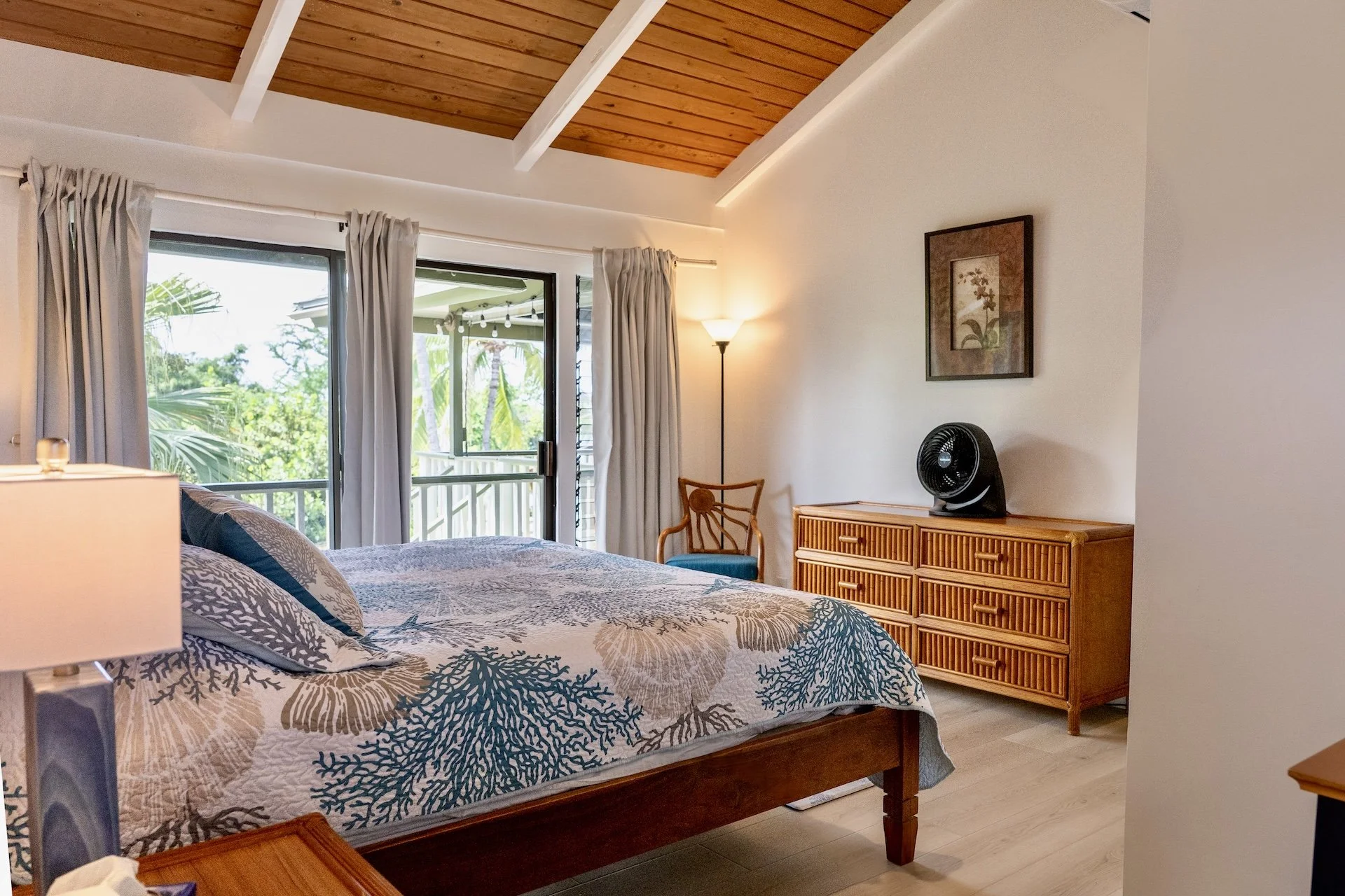 A bedroom with a bed that has a coral-themed bedspread, a wooden dresser with a small fan on top, and a picture of flowers on the wall. There are sliding glass doors with curtains leading to a balcony with greenery outside.