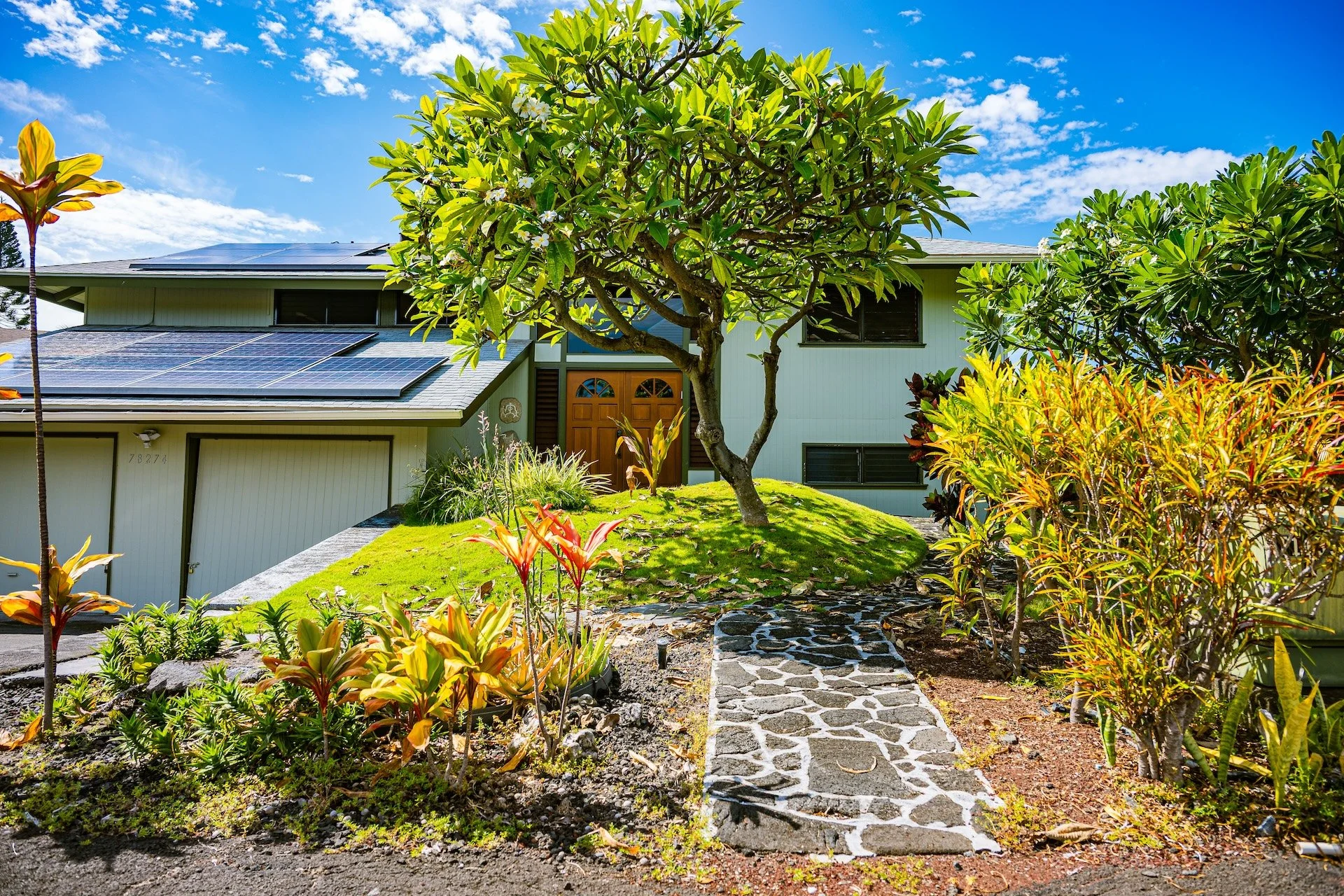 A house with solar panels on the roof, surrounded by lush green plants, trees, and a stone pathway leading to the front door, under a bright blue sky with some scattered clouds.