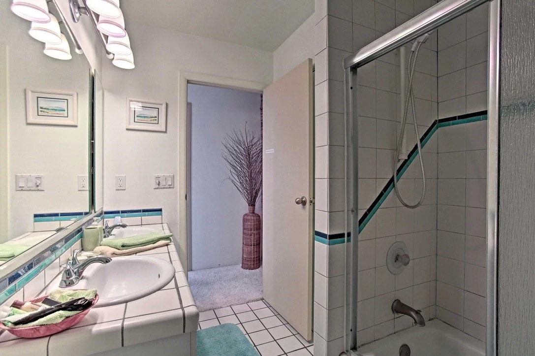 Bathroom with a white tiled vanity, green towels, a sink, a mirror, a shower with glass door, and a decorated hallway with a vase and plant.