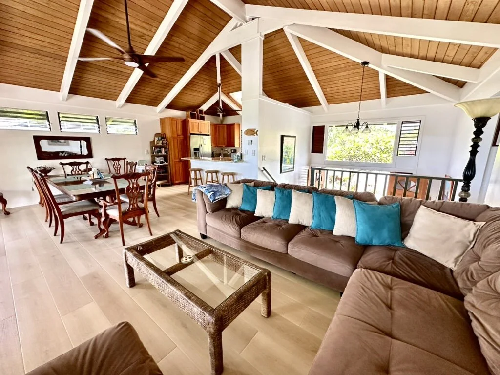 Open-concept living room, dining area, and kitchen in a house with vaulted wooden ceiling, ceiling fans, large windows, and a mixing of toasty brown and bright white decor.