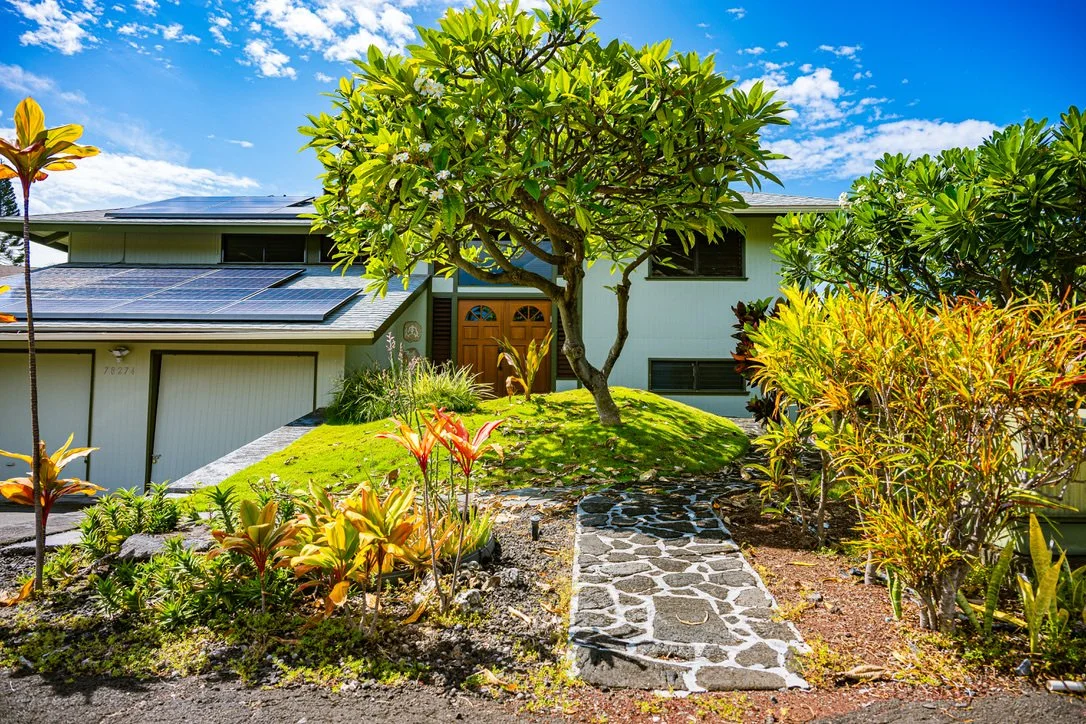 A house with solar panels on the roof, surrounded by a well-maintained garden with a variety of plants, a stone pathway, and a large tree in the front yard.