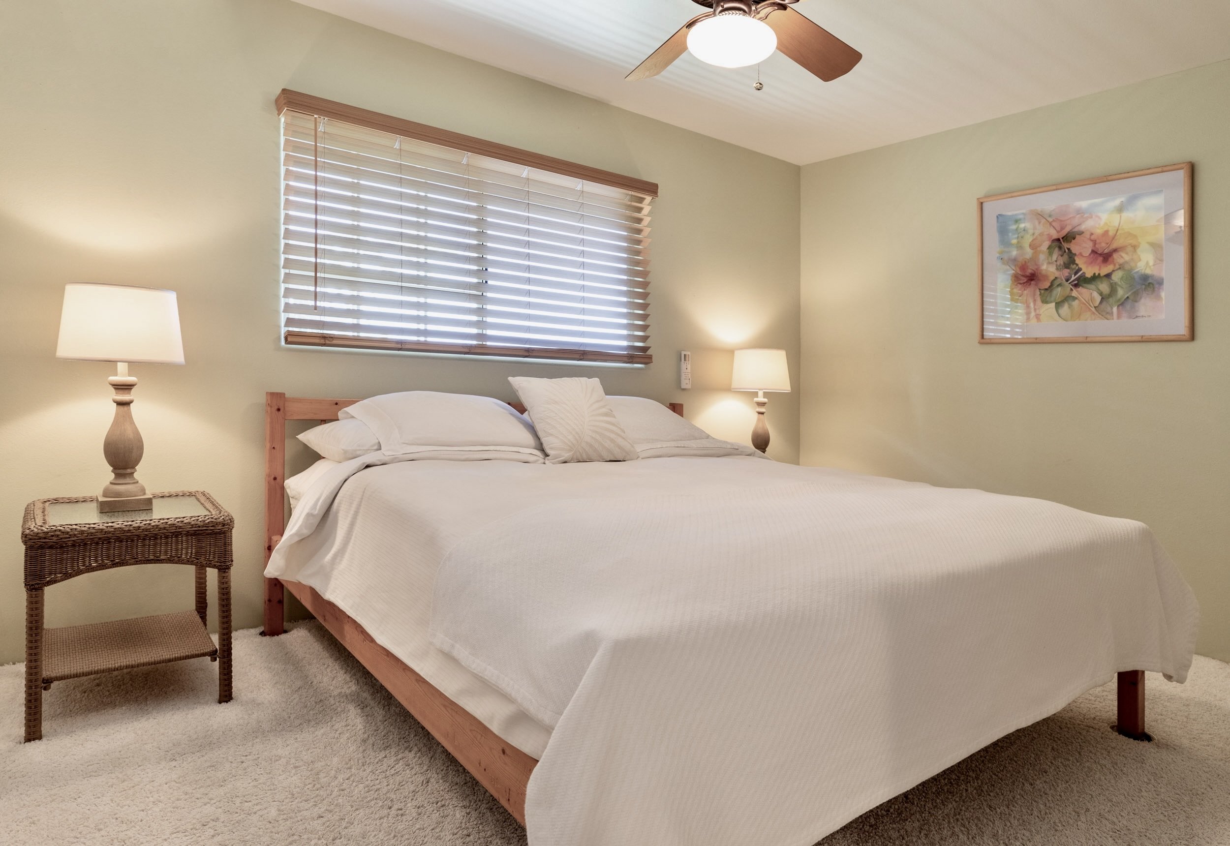 A bedroom with a wooden bed frame, white bedding, and pillows. Two nightstands with lamps are on each side of the bed. A window with wooden blinds is above the headboard, and a framed floral watercolor painting is on the wall. A ceiling fan with wood