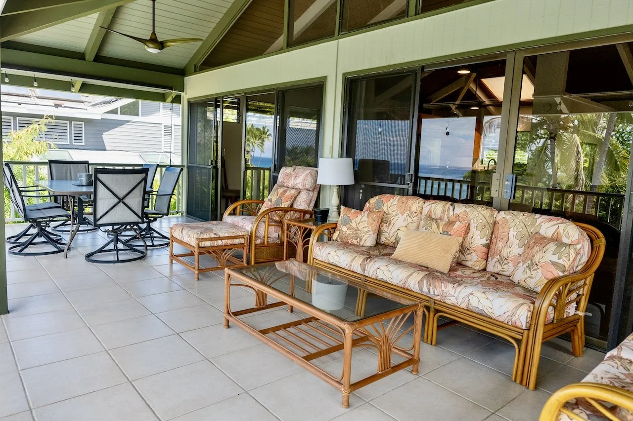 Sloped covered balcony with outdoor furniture, including a floral-patterned wicker sofa and chairs, a glass-top coffee table, and a dining table with chairs, overlooking greenery and water.