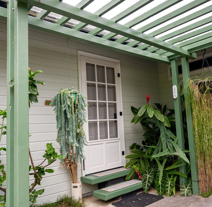 Close-up of a front porch with a white door, green wooden pergola, plants, and a small step.