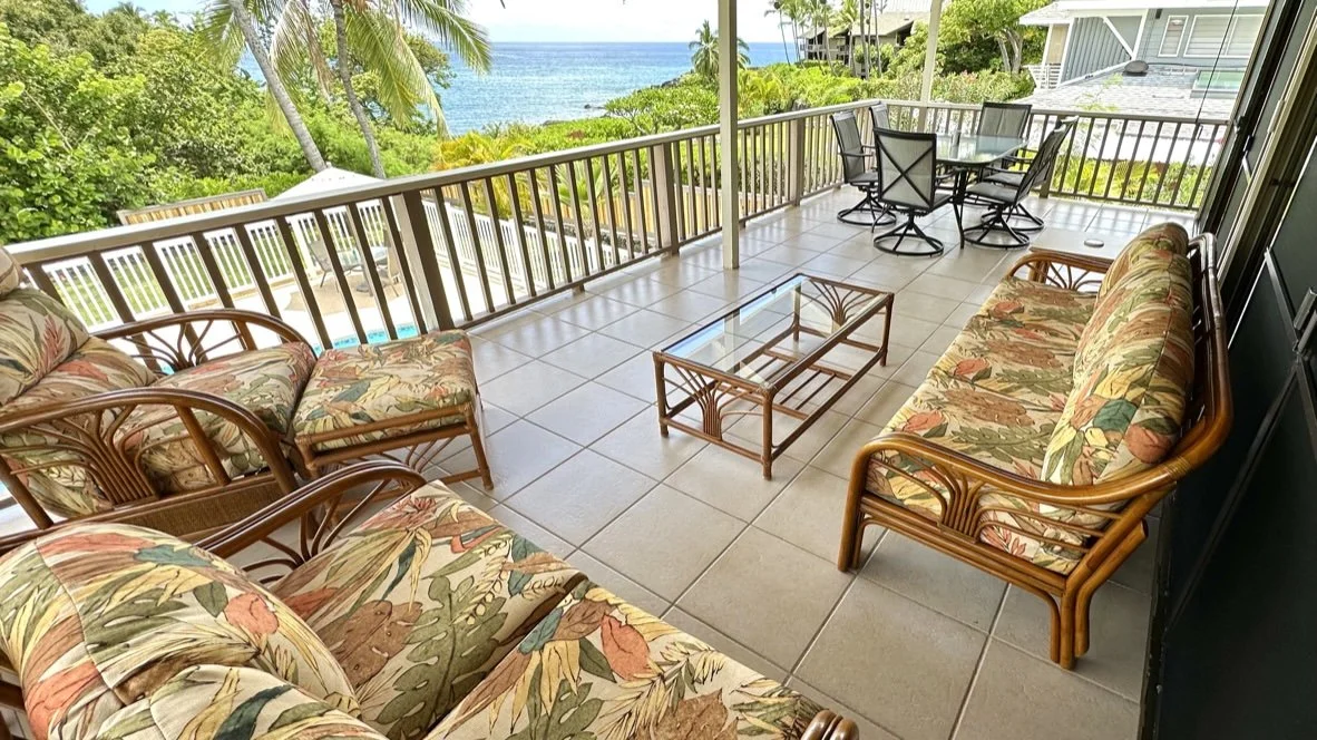 Covered patio with tropical furniture and a view of lush greenery and the ocean in the distance.