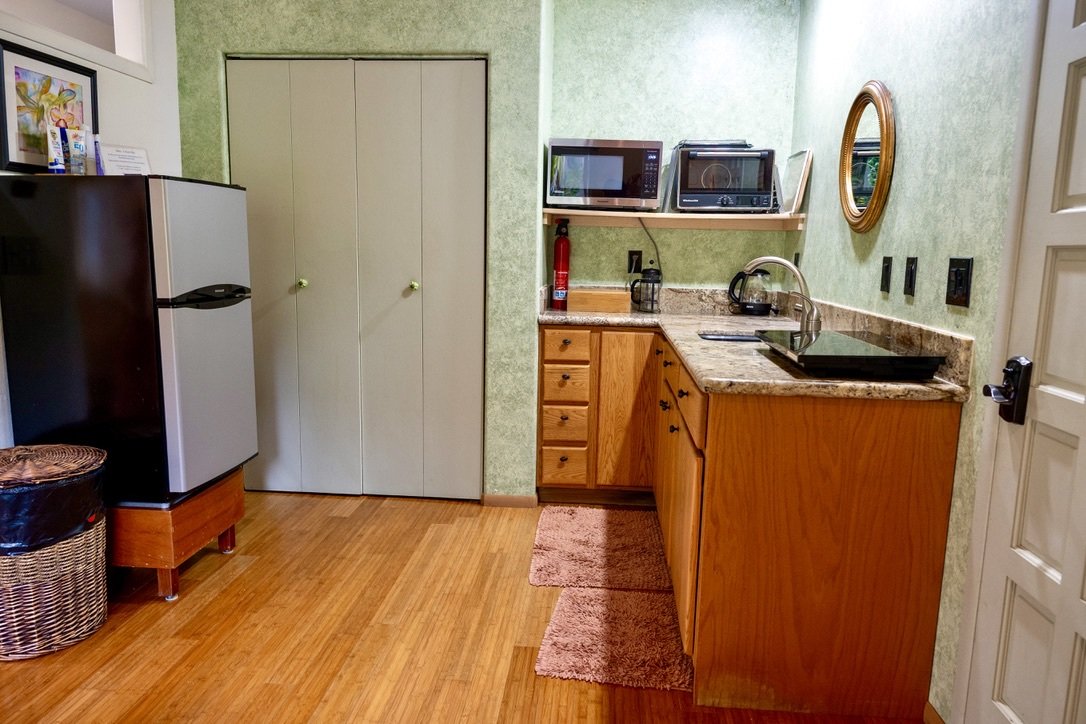 Compact kitchen with granite countertop, wooden cabinets, microwave, toaster, kettle, fire extinguisher, and small mirror on green textured wall, hardwood floor, and curtains in the background.