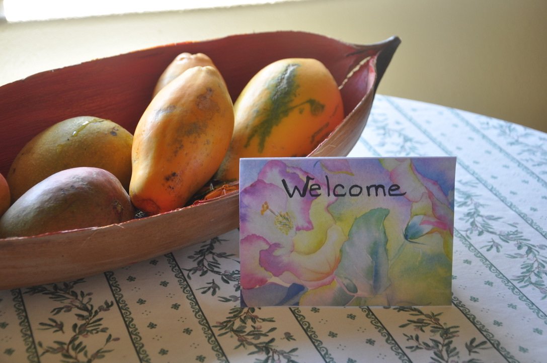 A wooden bowl with mangoes on a table with a floral tablecloth. A colorful welcome card with watercolor flower design is in front of the bowl.
