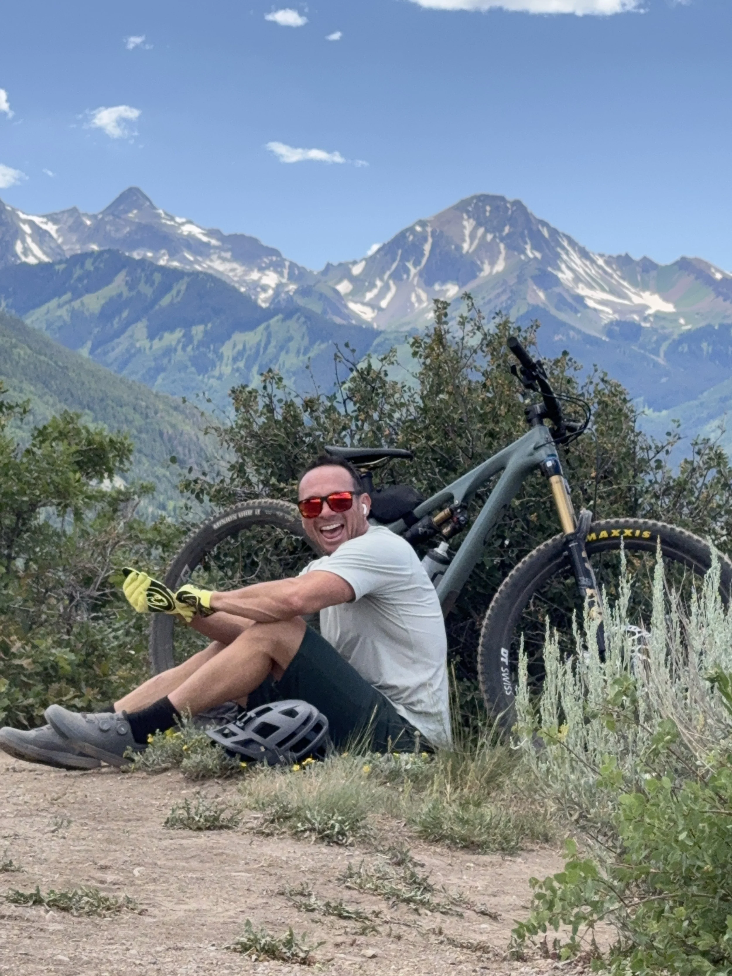 Luke Jasso sitting in the foreground, smiling, with a mountain bike gear, in a natural setting with mountains in the background landscape.