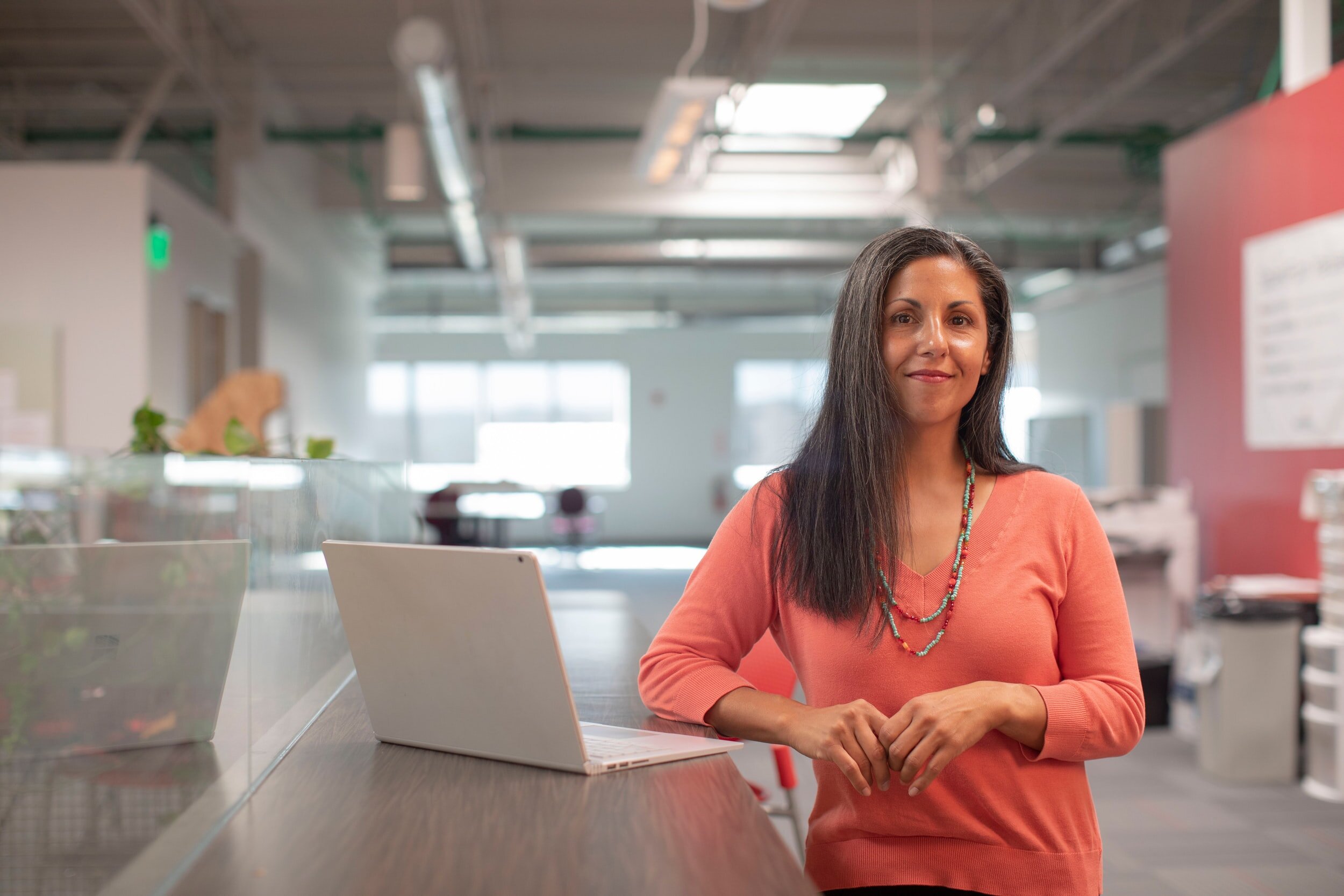 Woman in coral sweater standing by a table with a laptop in a modern office environment.
