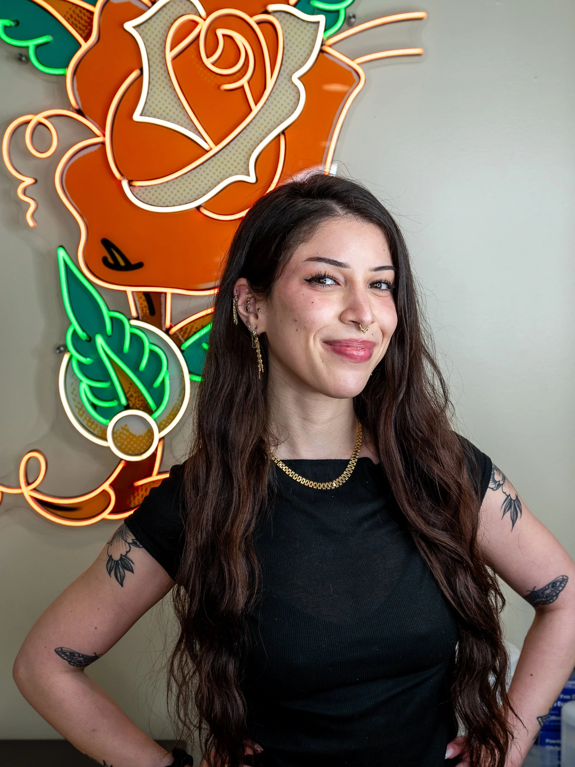 A woman with long, wavy brown hair, dressed in a black shirt, smiles confidently with her hands on her hips in front of a colorful neon sign of a stylized rose with green leaves.