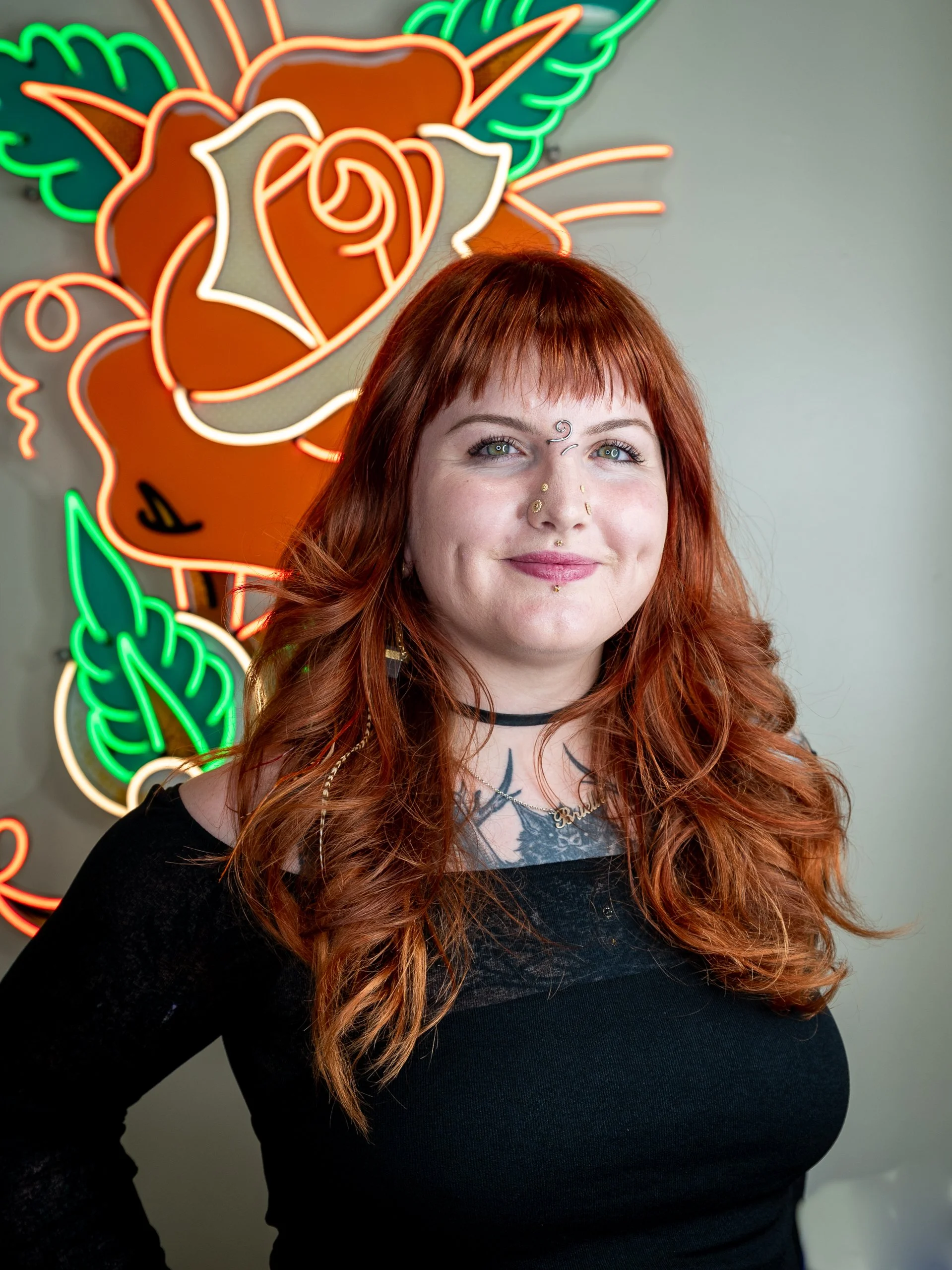A woman with long red hair, tattoos, and facial piercings smiling in front of a neon rose sign.