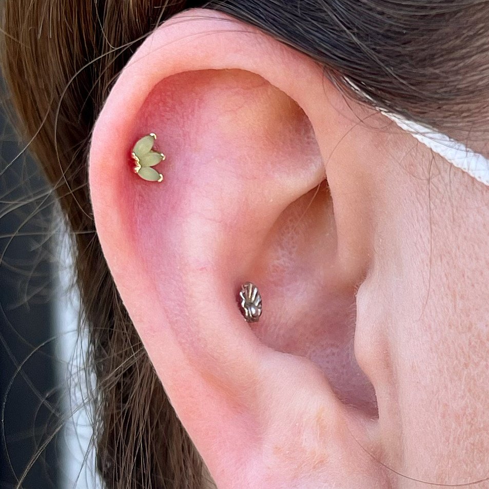 Close-up of a human ear with two cartilage piercings, featuring a green flower stud and a circular silver stud.