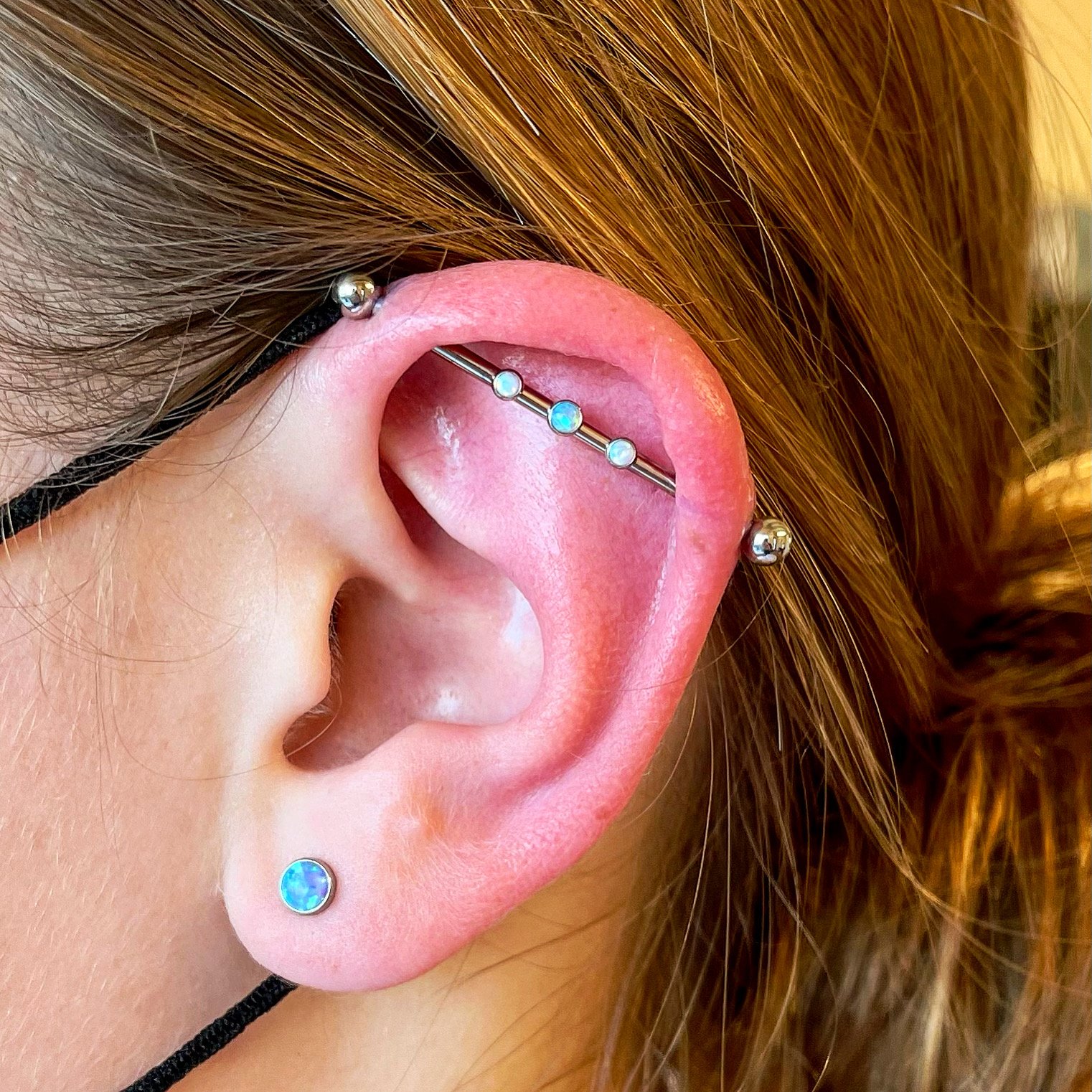 Close-up of an ear with a double cartilage piercing featuring a straight barbell and gemstone studs, along with a small stud in the earlobe.