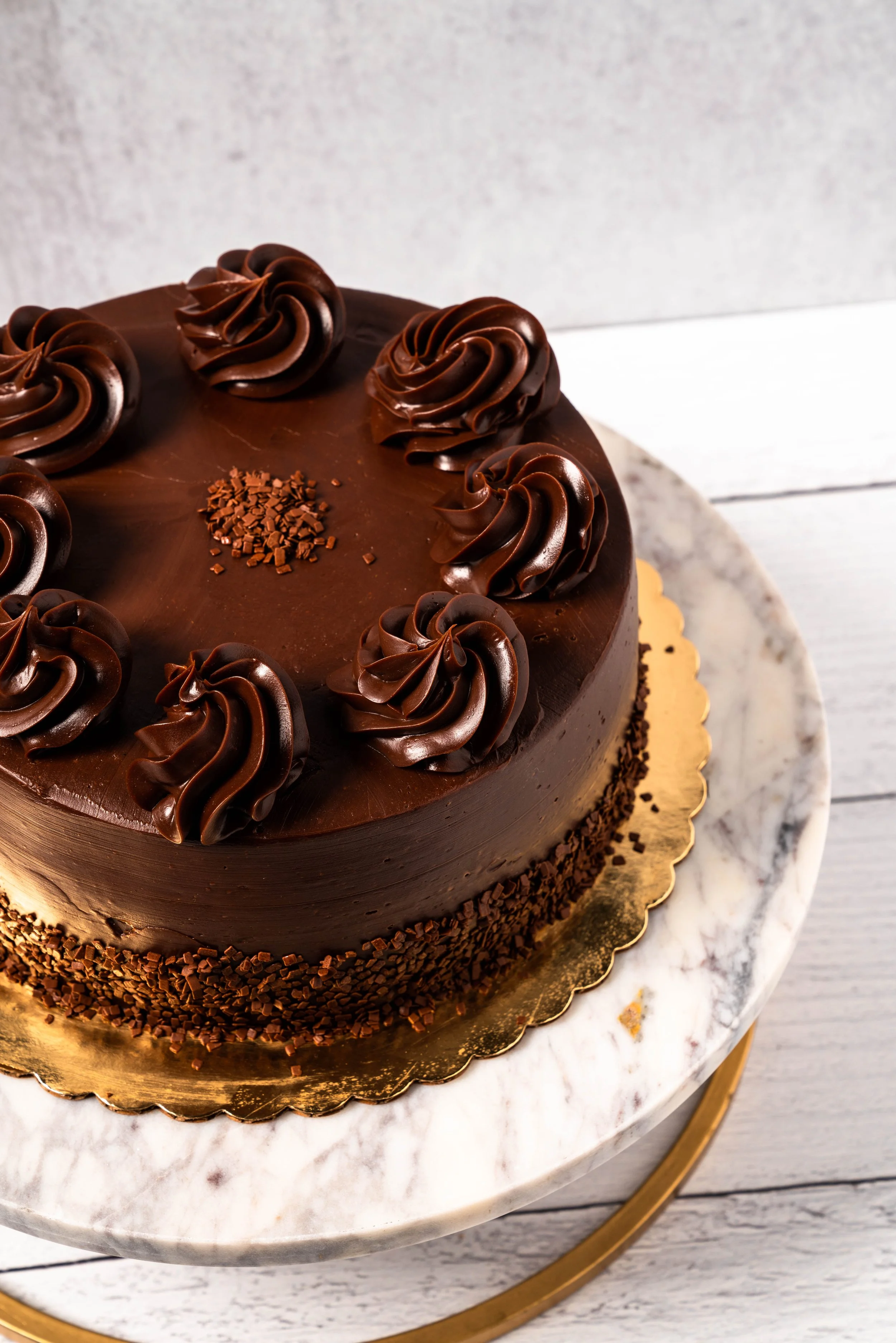 Chocolate cake with piped chocolate frosting rosettes on top, chocolate shavings around the sides, and a small pile of chocolate sprinkles in the center, placed on a marble serving board.
