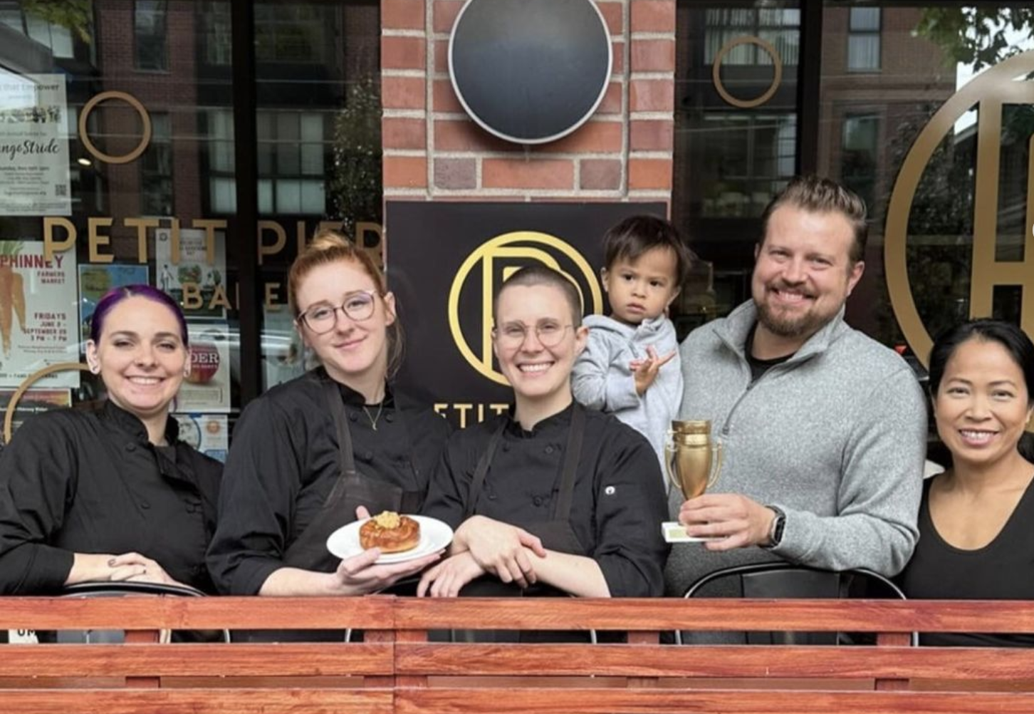 Group of six people, four women and two men, standing outside a restaurant, smiling. Two women and one man wear black chef jackets, holding a plate with a pastry. The man is holding a small trophy, and a child is sitting on the man's shoulder. The restaurant window has decorative lettering and posters.