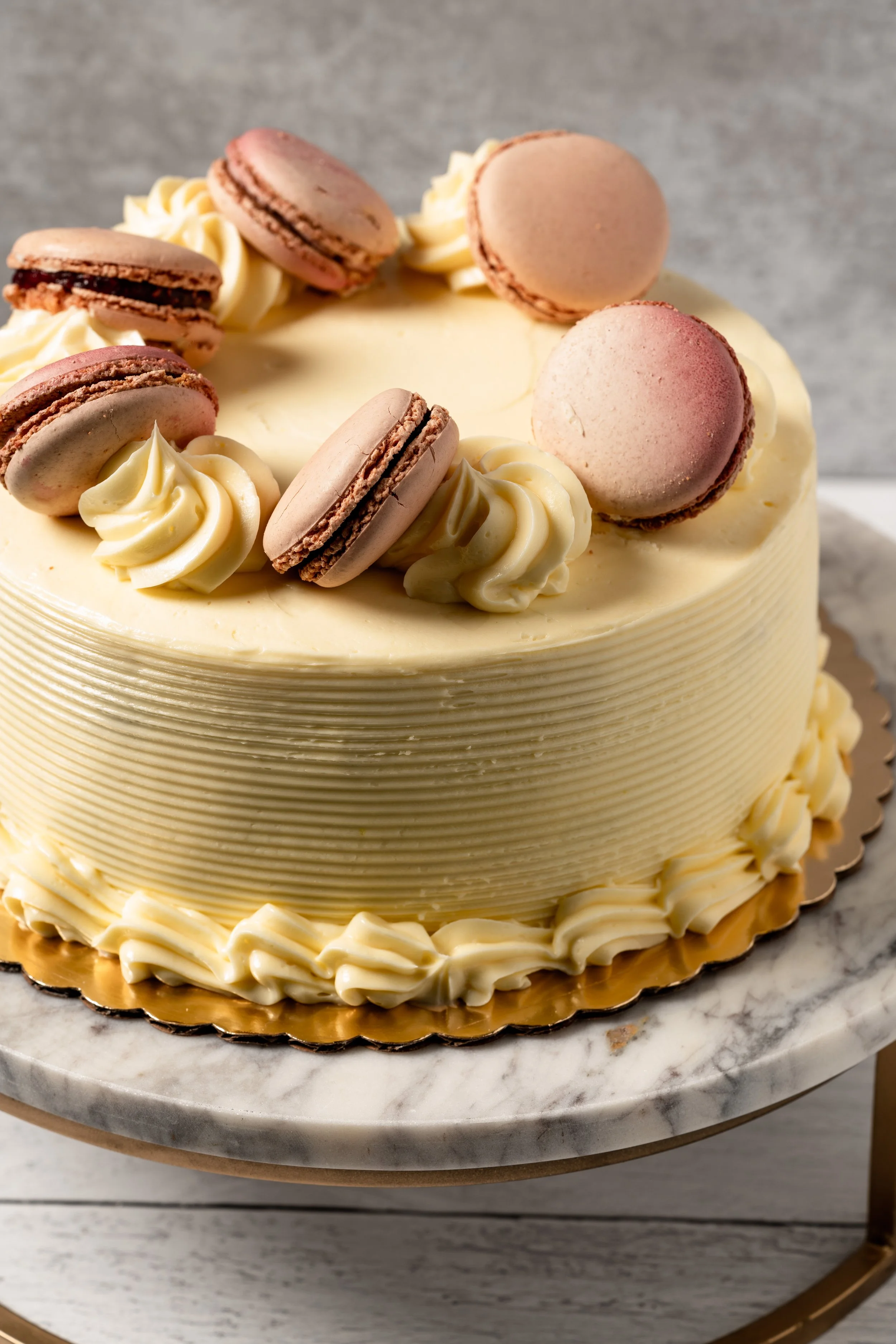 A round white cake decorated with pink macarons and white cream rosettes on top, placed on a marble cake stand.