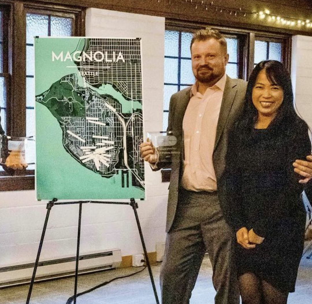 A man and woman standing indoors, smiling at the camera, with a green and black map titled 'Magnolia' on a tripod behind them.