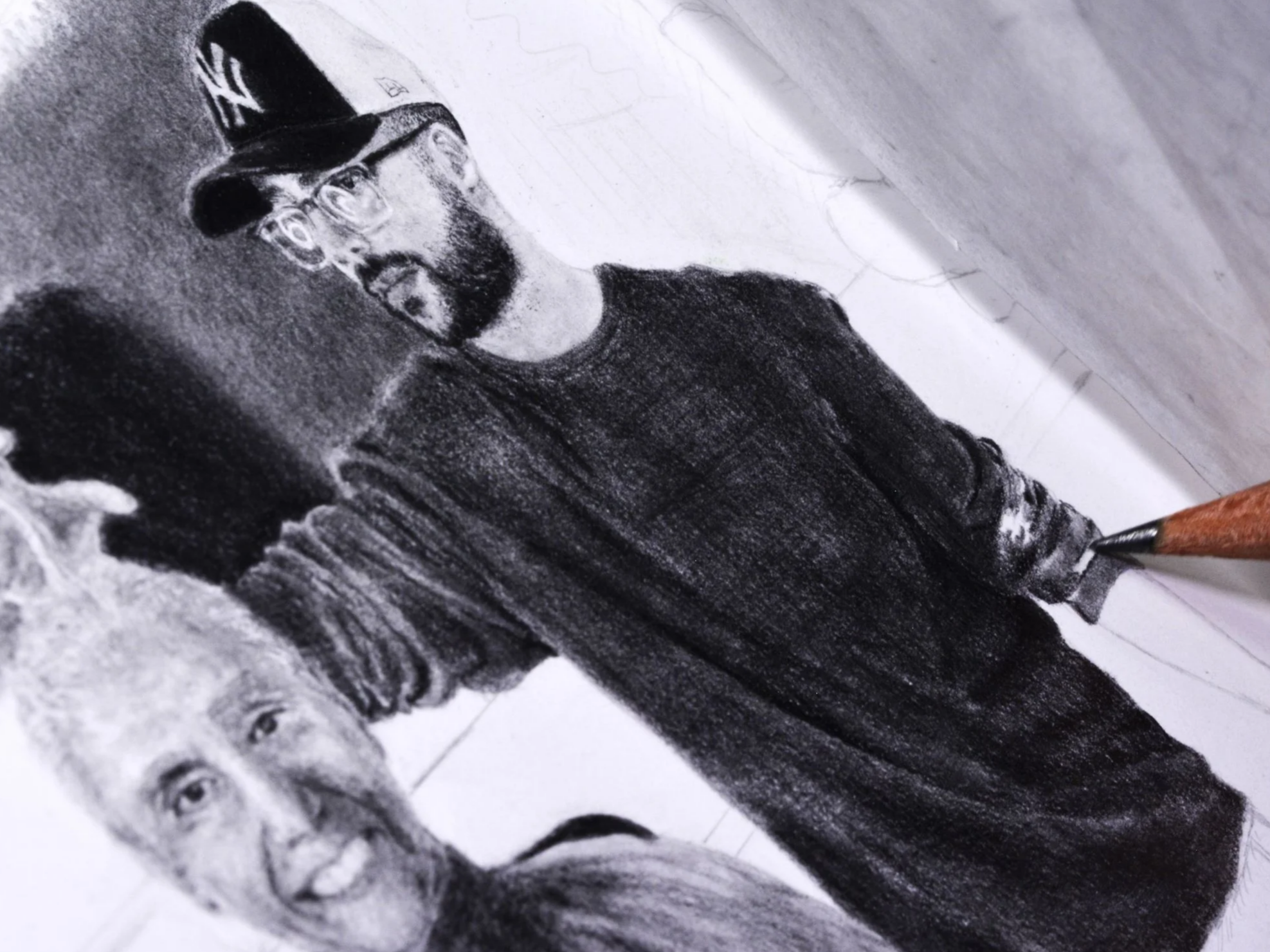 Photograph of a detailed black and white pencil sketch of two people, one with glasses and a baseball cap, and the other with curly hair, on white paper.