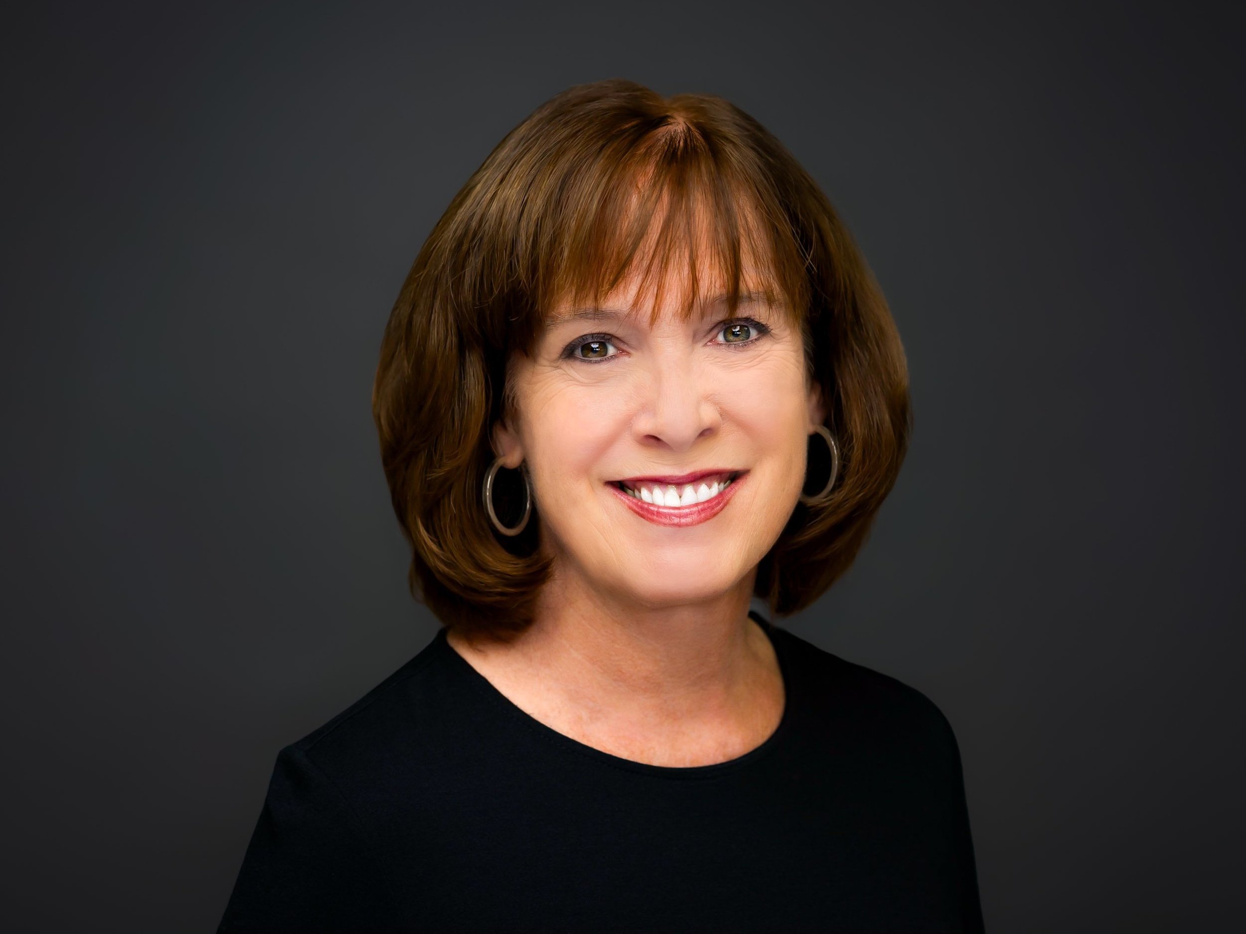 Professional headshot of a smiling woman with shoulder-length brown hair, wearing earrings and a black top against a dark gray background.