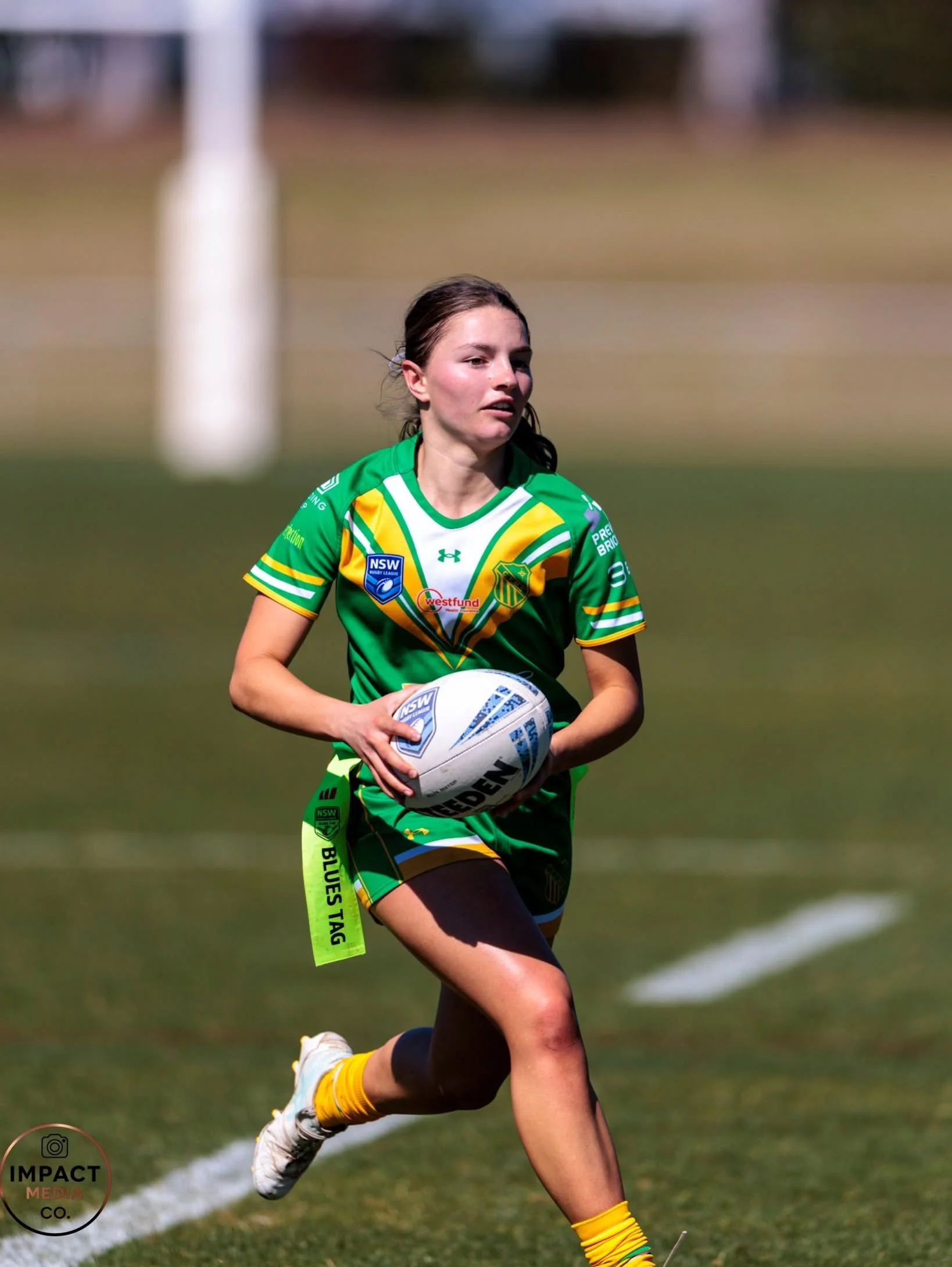Female rugby league player in green and yellow runs with the ball on a grass field.