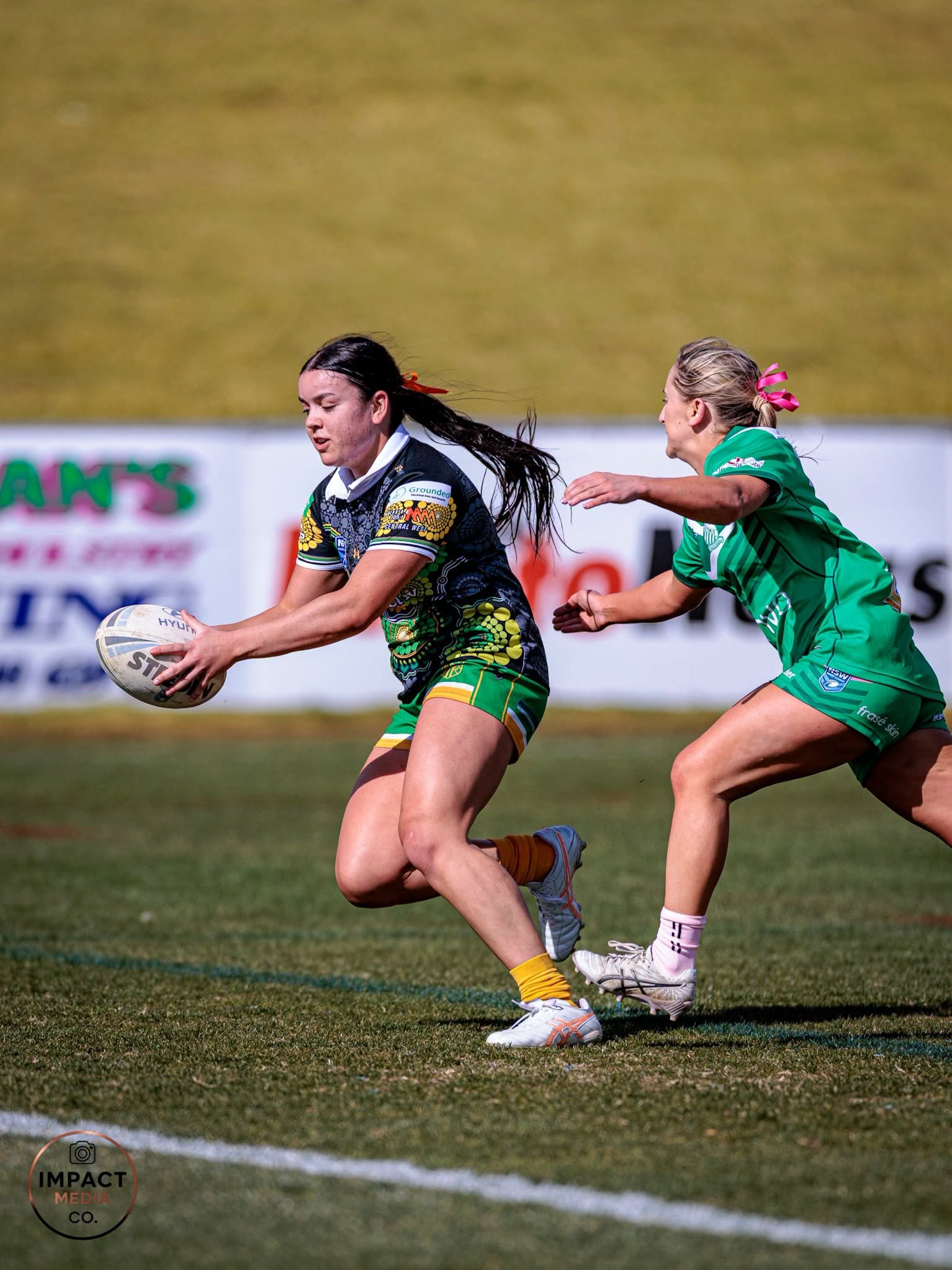 Two female rugby league players chase the ball carrier on field.